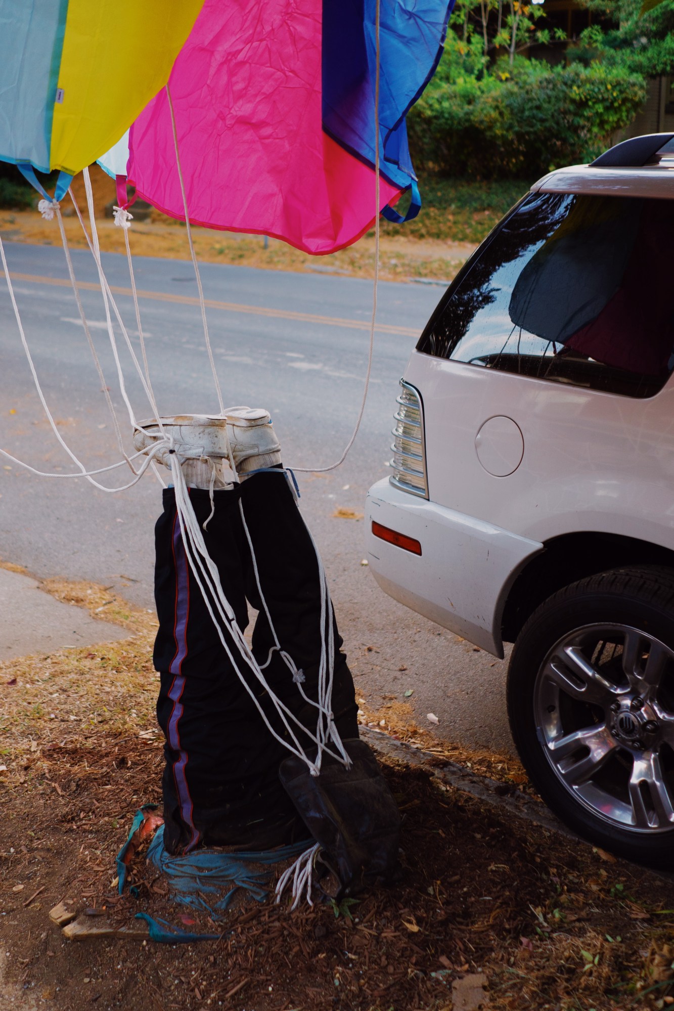 a figure half-hidden in the ground with balloons tied to their legs beside a white car