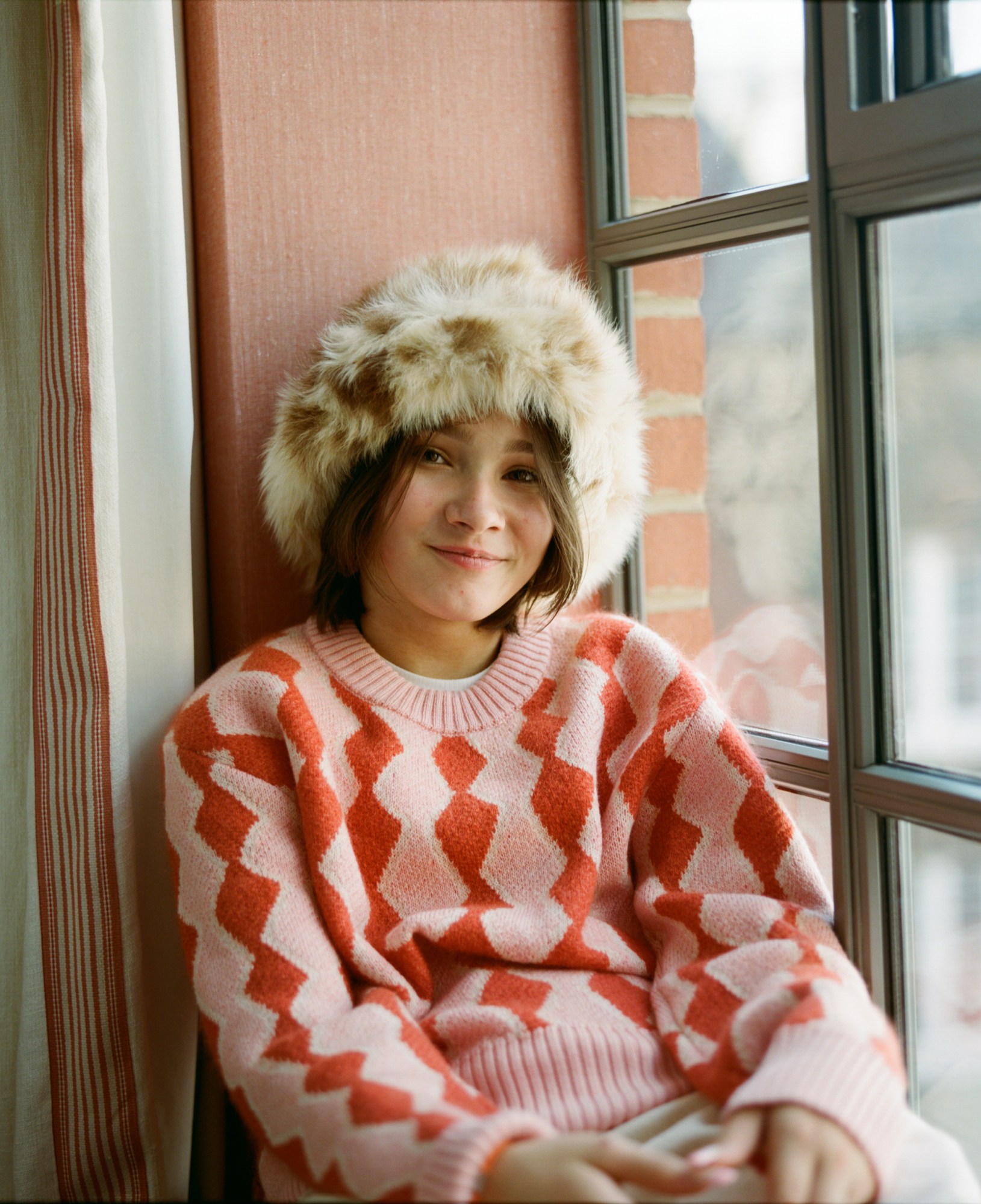 frankie sits on the windowsill wearing a furry hat and pink and red jumper