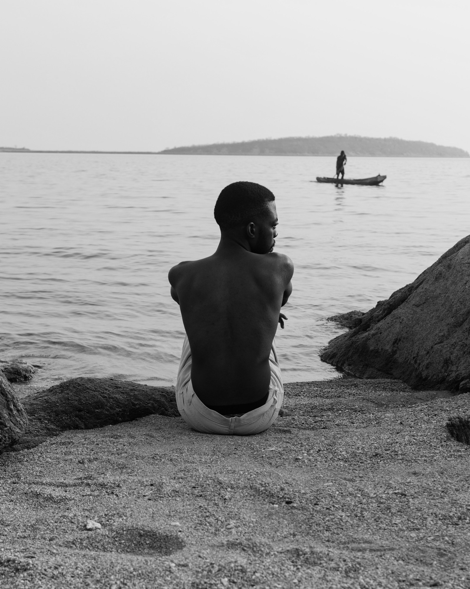 black-and-white image of a topless man sitting on the beach, shot from behind