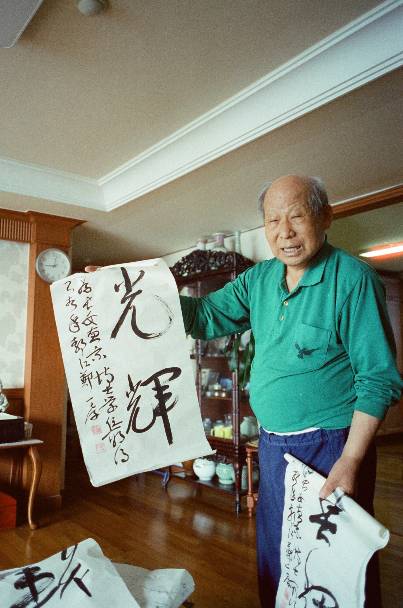 an old man shows off calligraphy wearing a green long-sleeved top and blue jeans