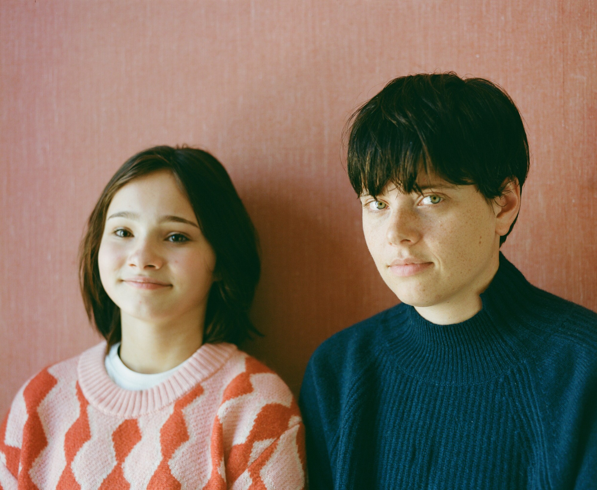 frankie and charlotte stand side by side against a pink backdrop