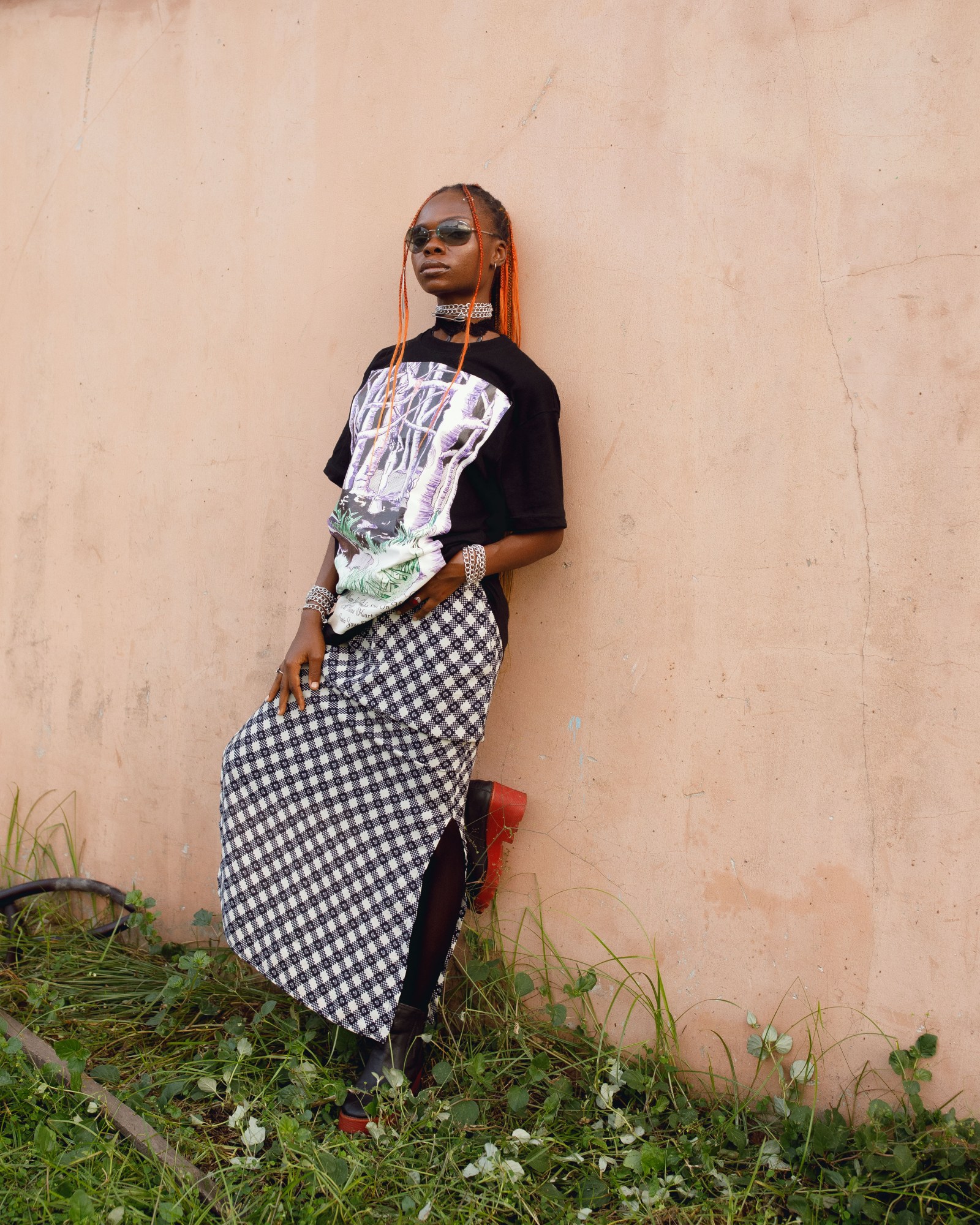 A woman with orange braids stood against a wall in a baggy printed t-shirt and a plaid skirt