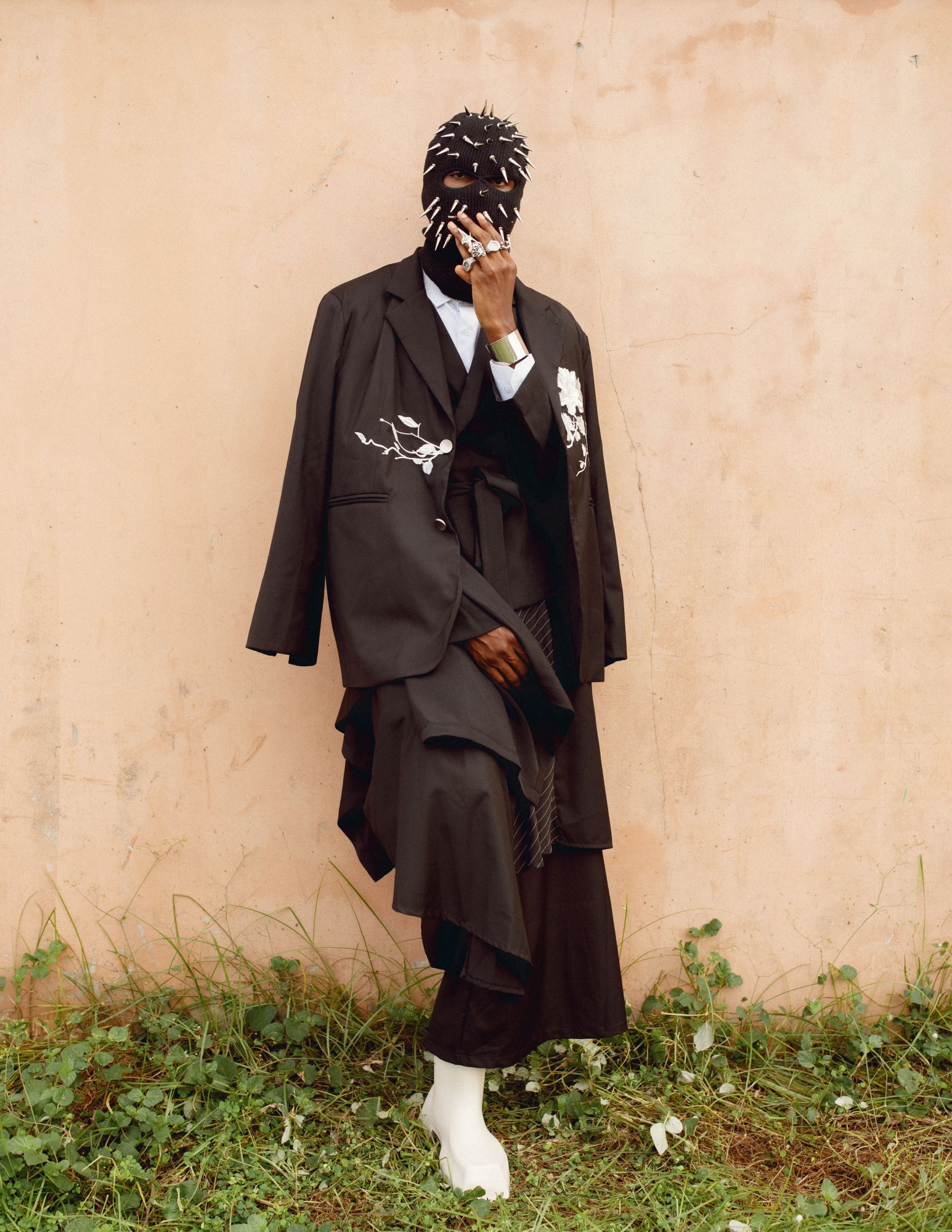 A person stood against a wall wearing a spiky mask and layered tailoring