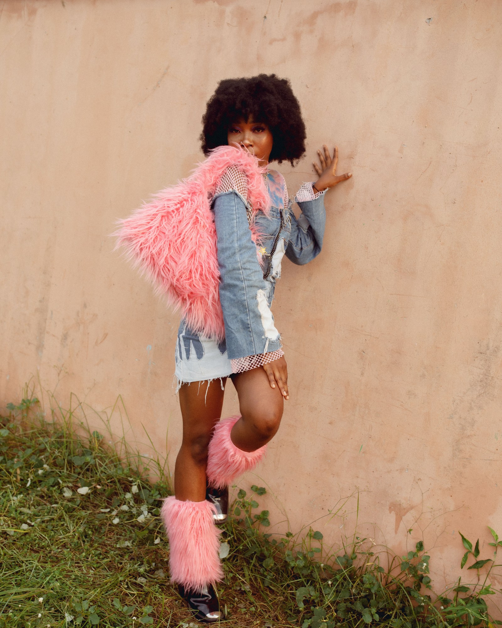 A woman stood against a wall in fluffy pink legwarmers and a bag, and a denim dress.