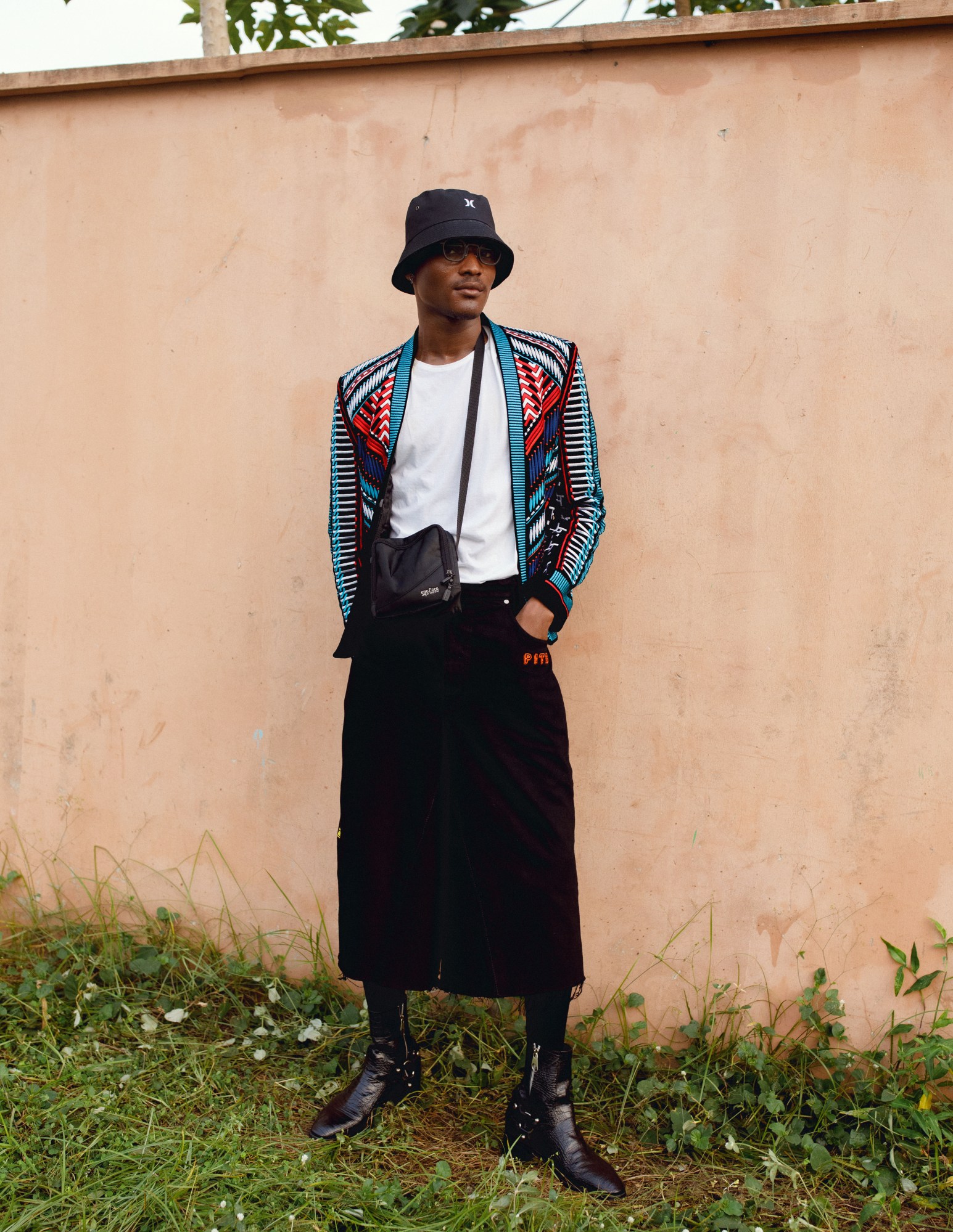 A man stood against a wall in leather ankle boots, a black skirt and a colourful jacket