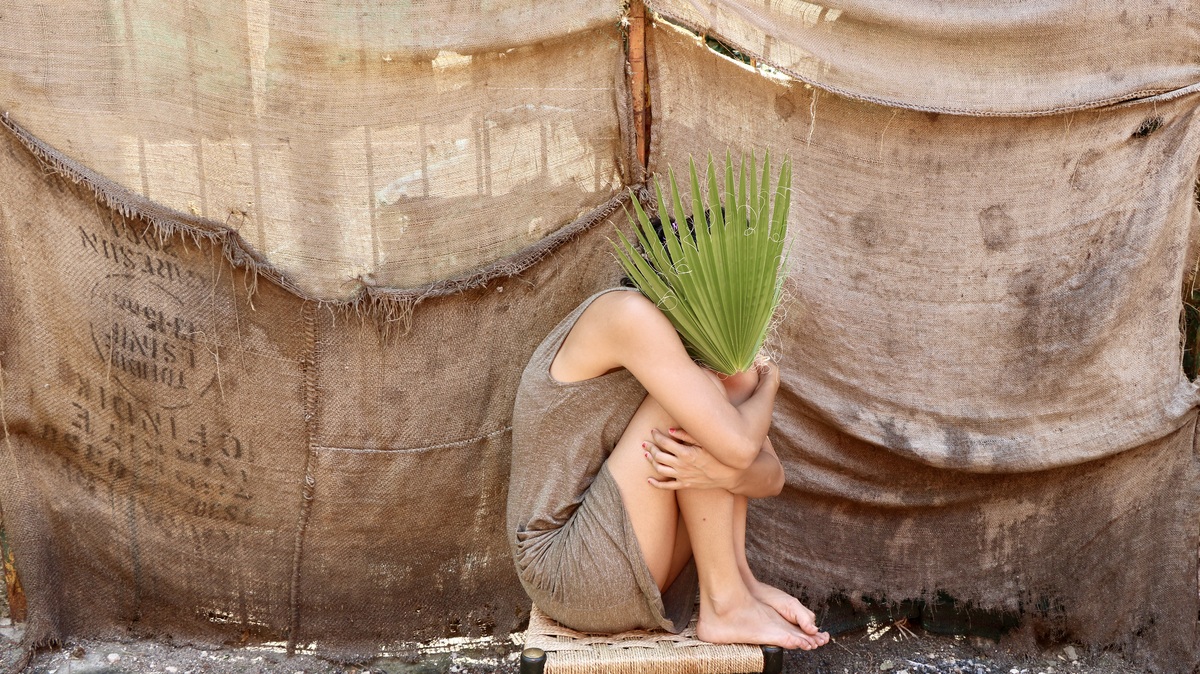 a woman in a brown dress sits huddled, hiding her face with a palm leaf