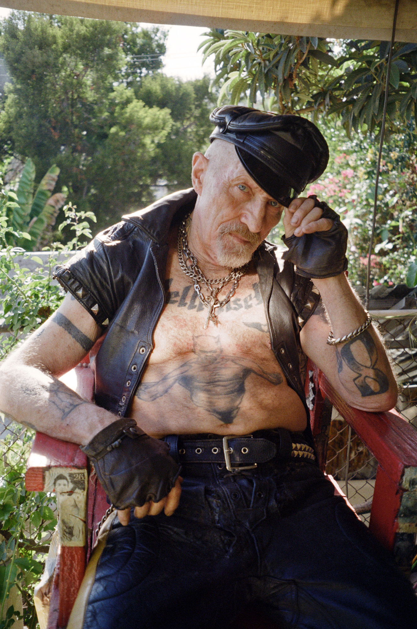 durk dehner wearing leather vest and gloves posing with tattoos and silver chains in a red chair