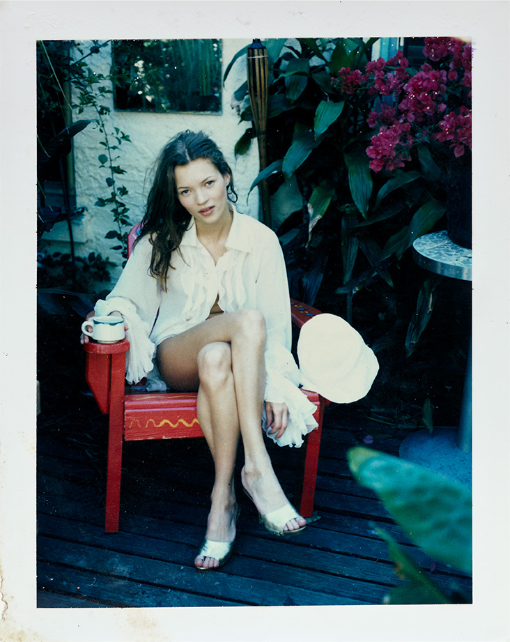 a young kate moss wearing a floaty white shirt sits in a garden with a coffee mug