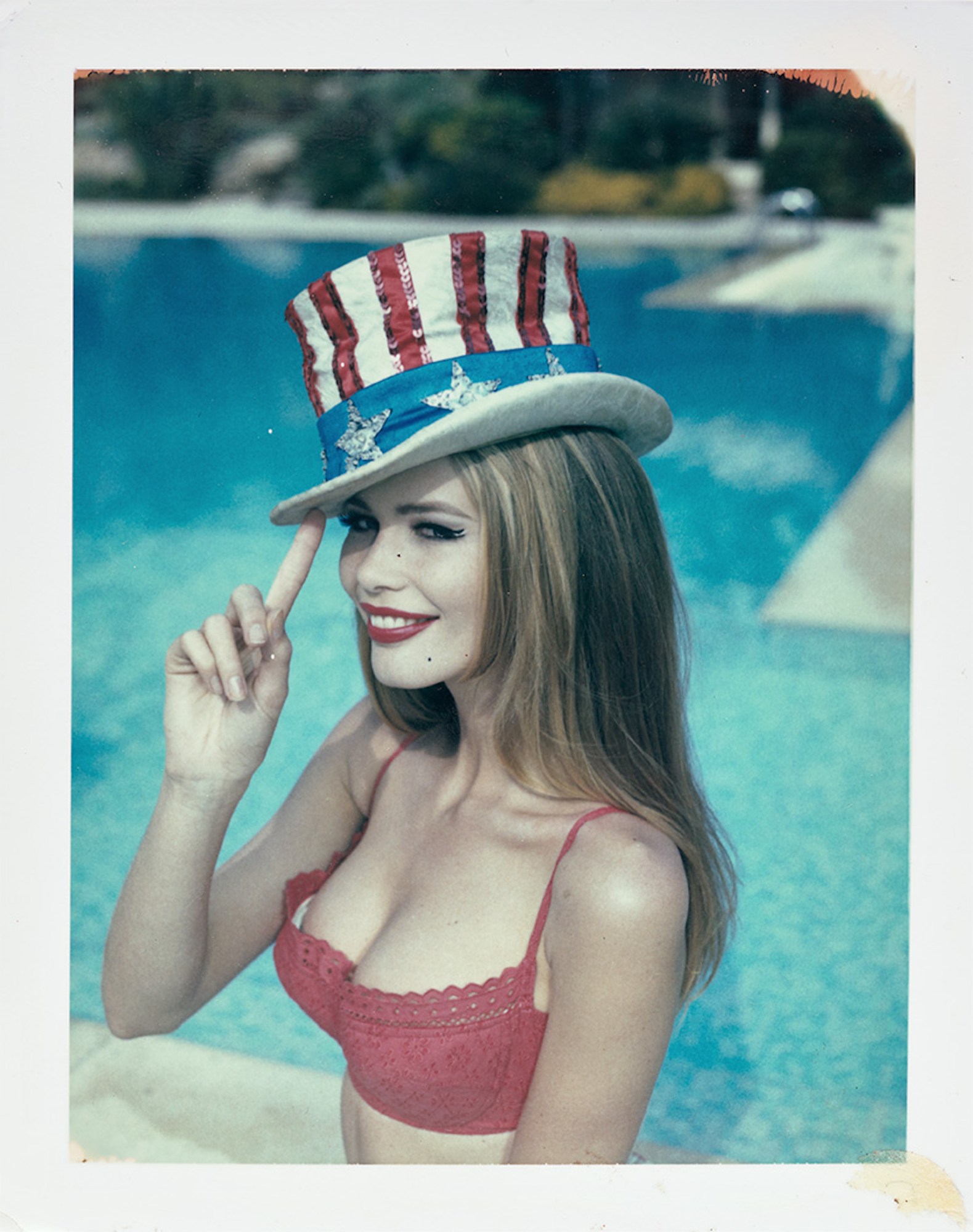 a young claudia schiffer sits by a pool wearing a red plunge bra and an uncle sam style US flag hat