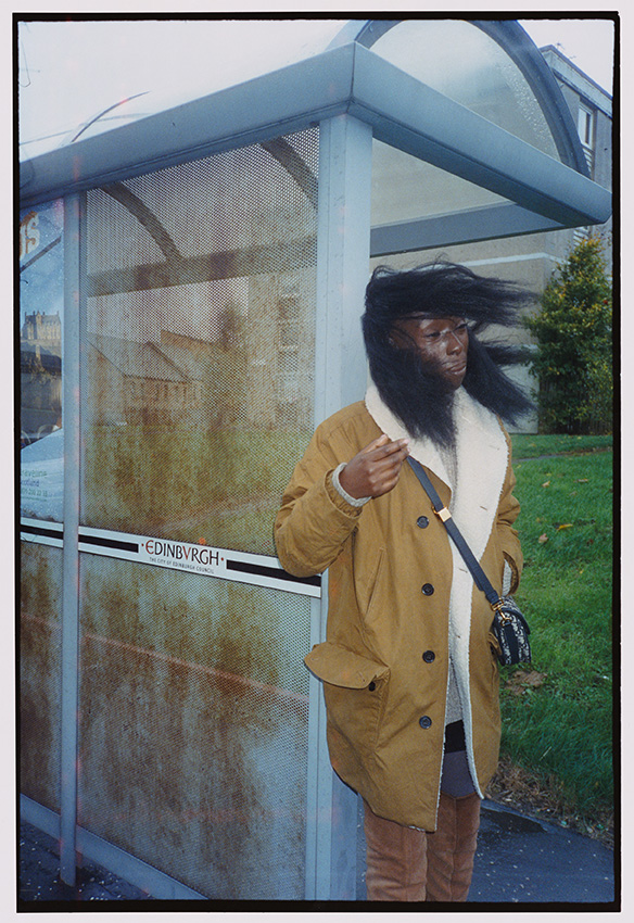 A woman in a brown coat stands by a bus stop as a gust of wind blows through her hair.