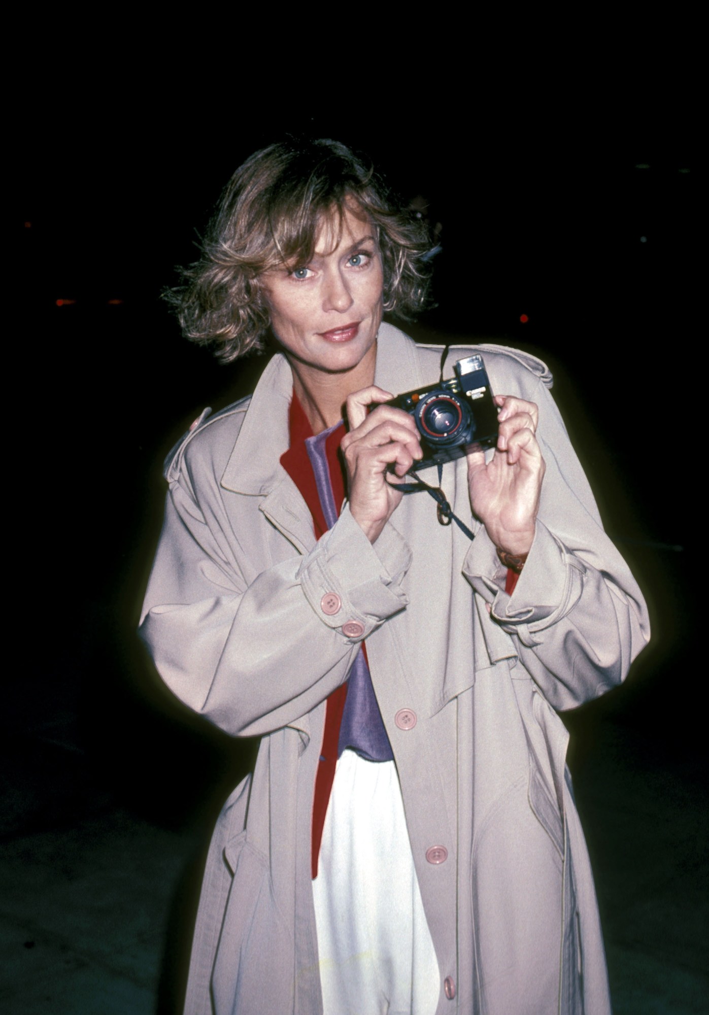 lauren hutton wearing a trench coat holding up a canon camera 1983