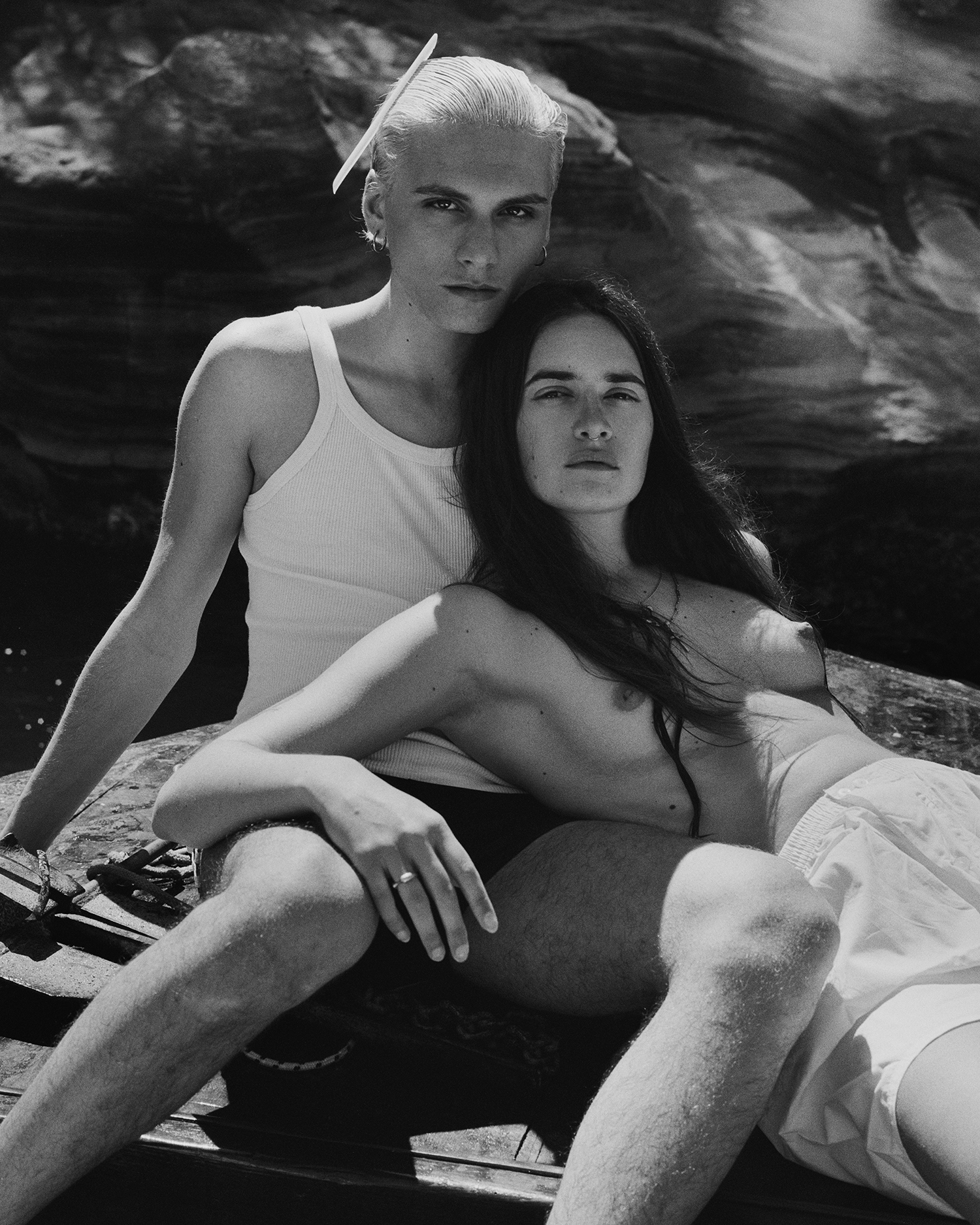 a couple recline together on a beach; one of them, a woman, is topless