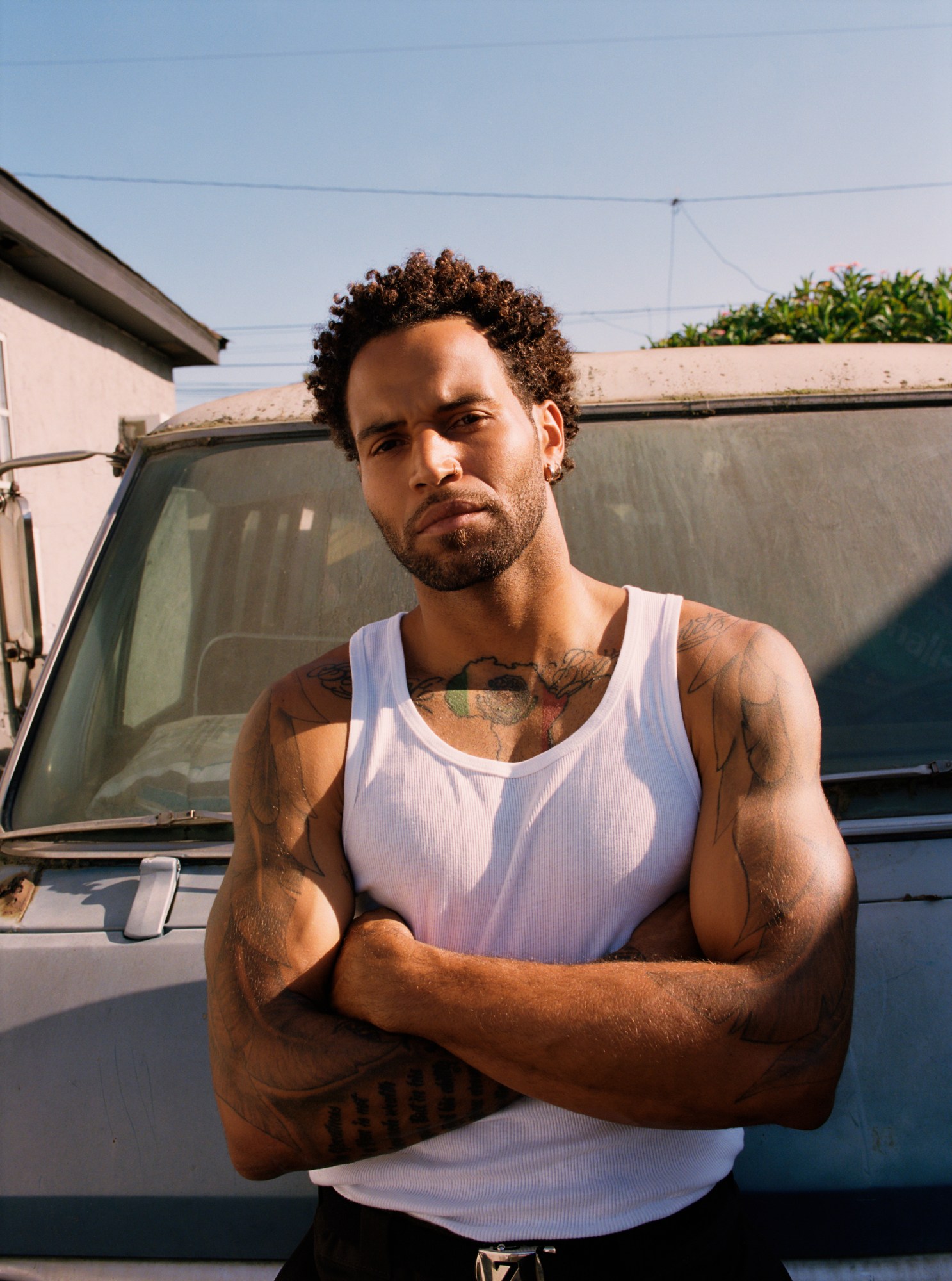 a man in a white tank top with arms crossed displaying his tattoos