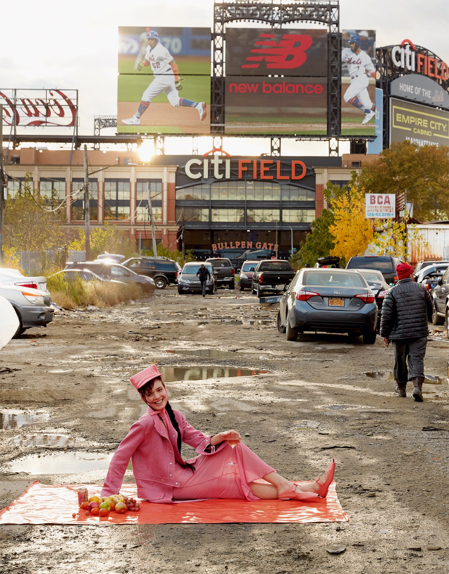 Photograph by Roe Ethridge of a woman in all pink sitting on a matt in a muddy shopping centre car park with fruit and a drink, featured in the book American Polychronic