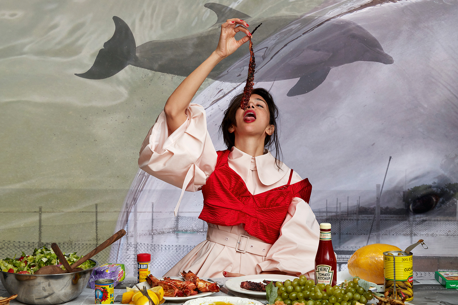 Photograph by Roe Ethridge of a woman in a dress and blouse top with food in front of her and eating meat while sitting in front of paintings of dolphins, featured in the book American Polychronic