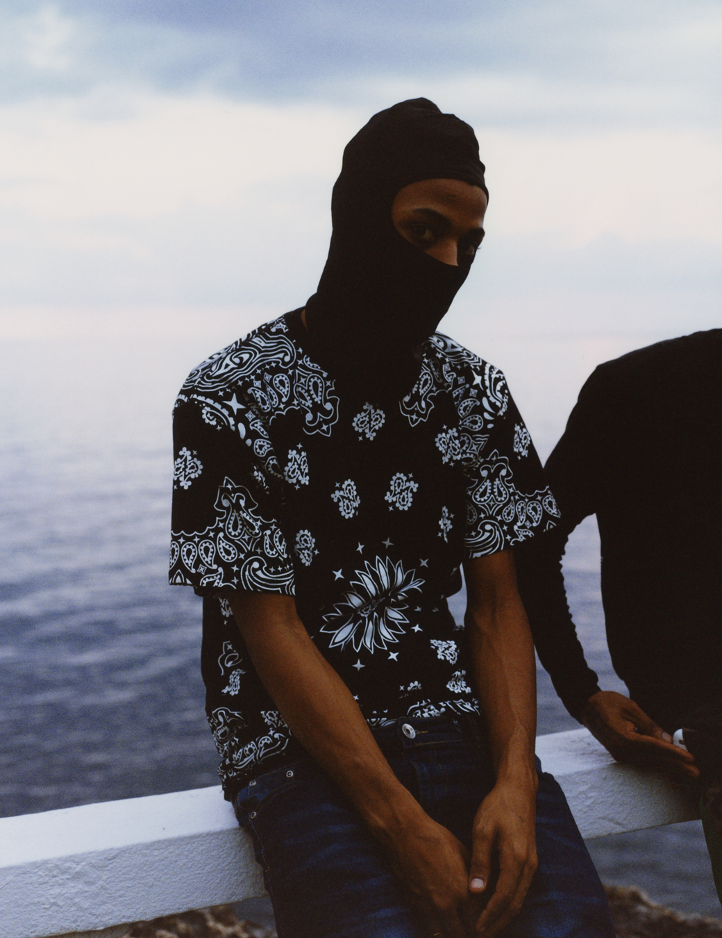 Bayka in a mask and shirt leaning against a rail looking over the ocean photographed by Jeano Edwards for i-D’s The Royalty Issue, no. 370, Winter 2022