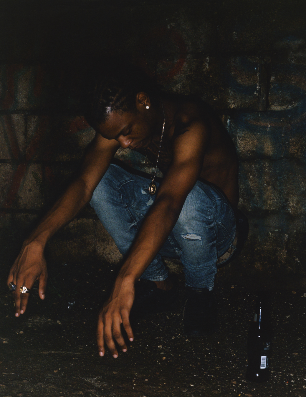 Pablo YG shirtless and crouching at night in front of a wall and next to a beer bottle photographed by Jeano Edwards for i-D’s The Royalty Issue, no. 370, Winter 2022