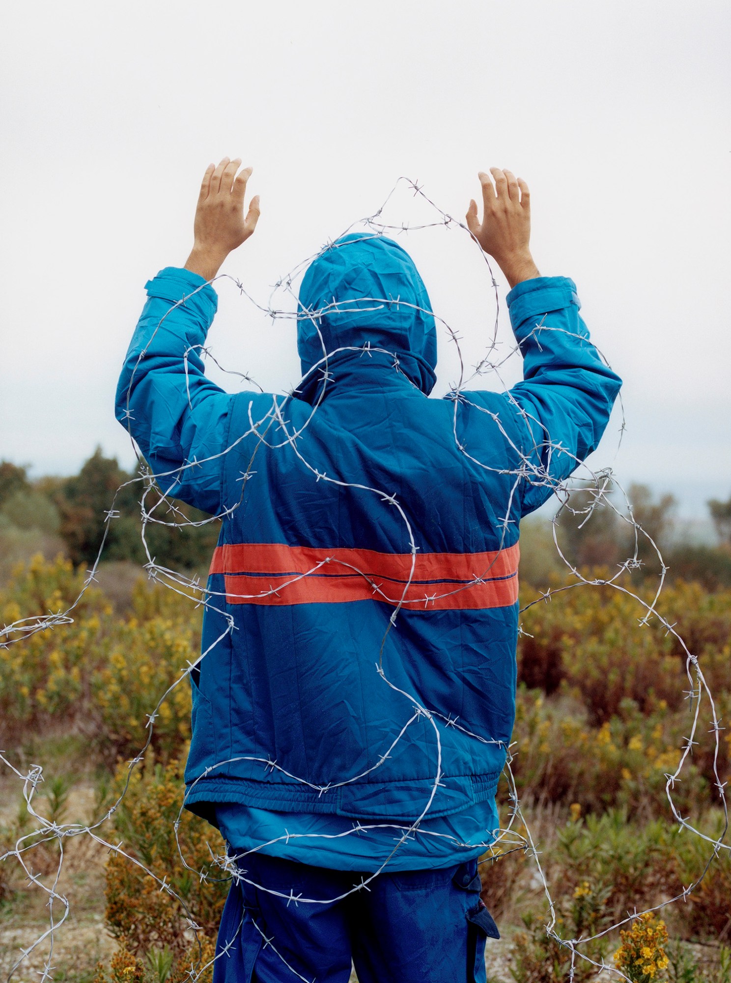 Photograph by Olgaç Bozalp of a boy in a blue coat and cargo pants from behind with his hands raised and wrapped in barbed wire standing in a field, featured in the book Leaving One for Another