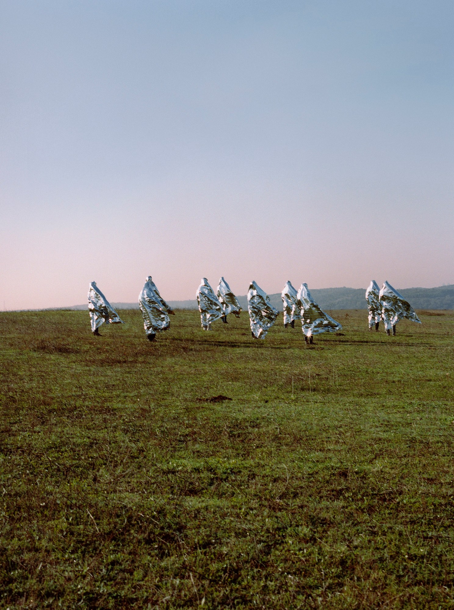 Photograph by Olgaç Bozalp of men in silver foil sheets walking along a grass field on a hill, featured in the book Leaving One for Another