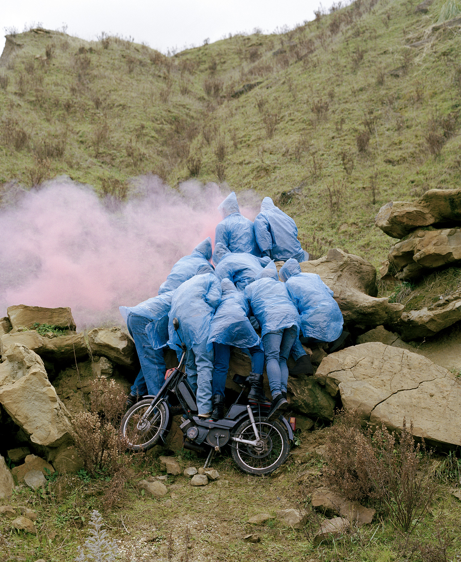 Photograph by Olgaç Bozalp of people in blue plastic coats and jeans crowded over rocks looking at a pink smoke, a motorbike in front of them, featured in the book Leaving One for Another