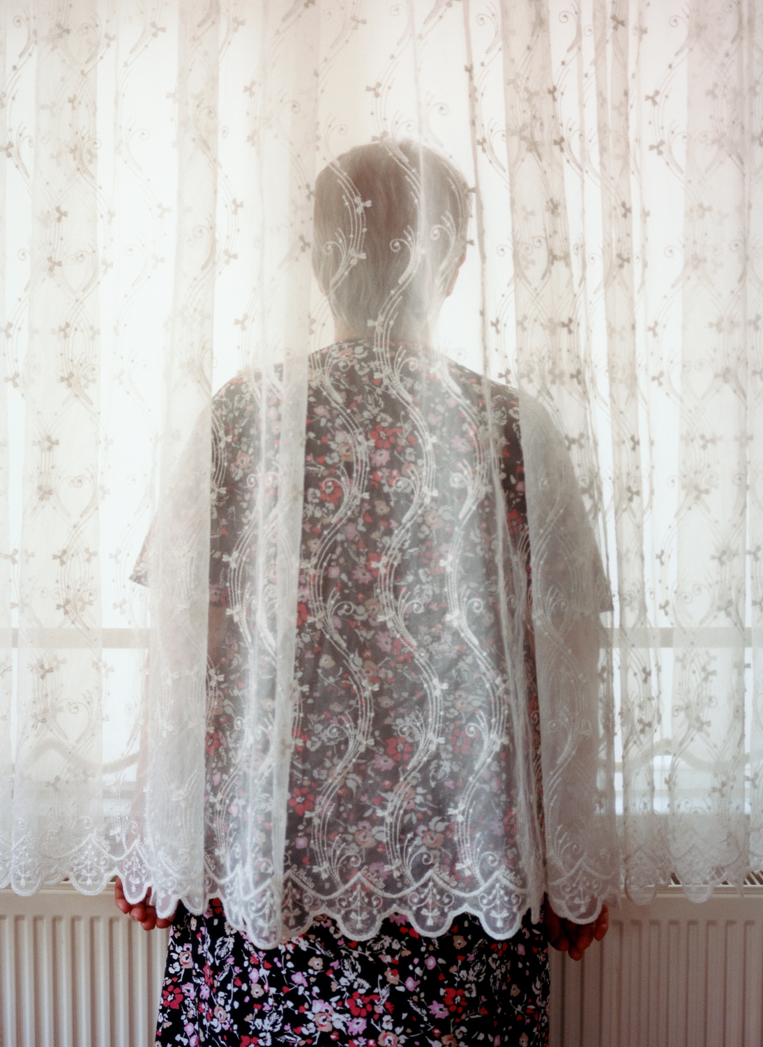 Photograph by Olgaç Bozalp of a woman in a floral dress standing under a net curtain looking out the window, featured in the book Leaving One for Another