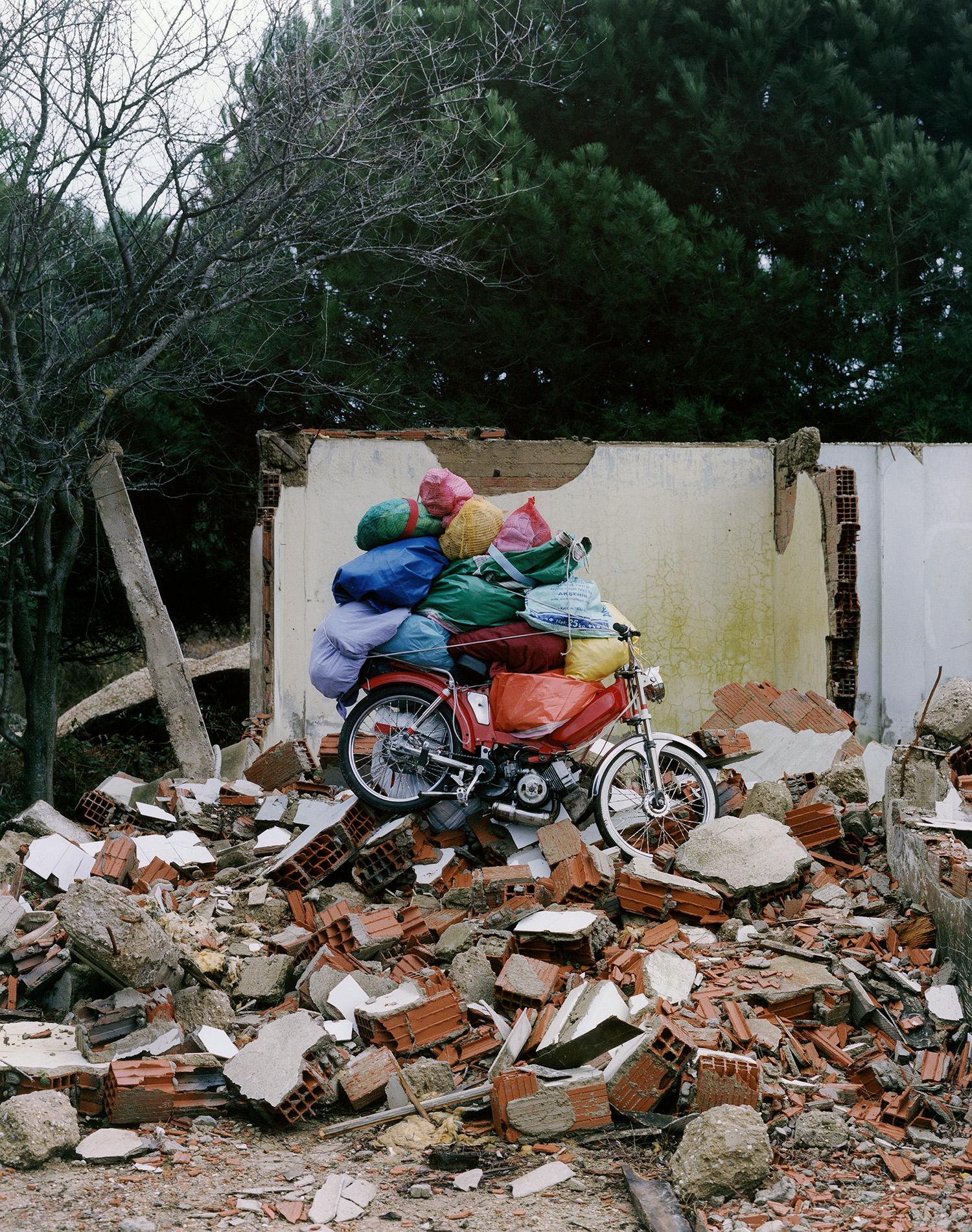 Photograph by Olgaç Bozalp of a motorbike sitting on rubble and loaded with colourful plastic bags, featured in the book Leaving One for Another