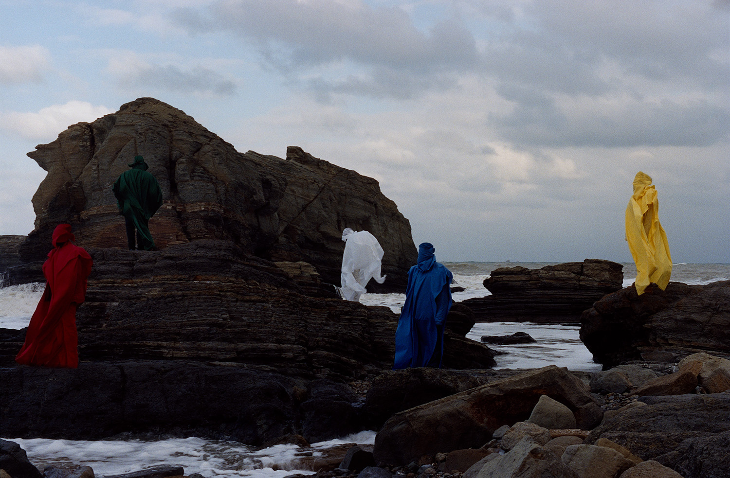 Photograph by Olgaç Bozalp of people in colourful burkas standing on rocks by a body of water, featured in the book Leaving One for Another