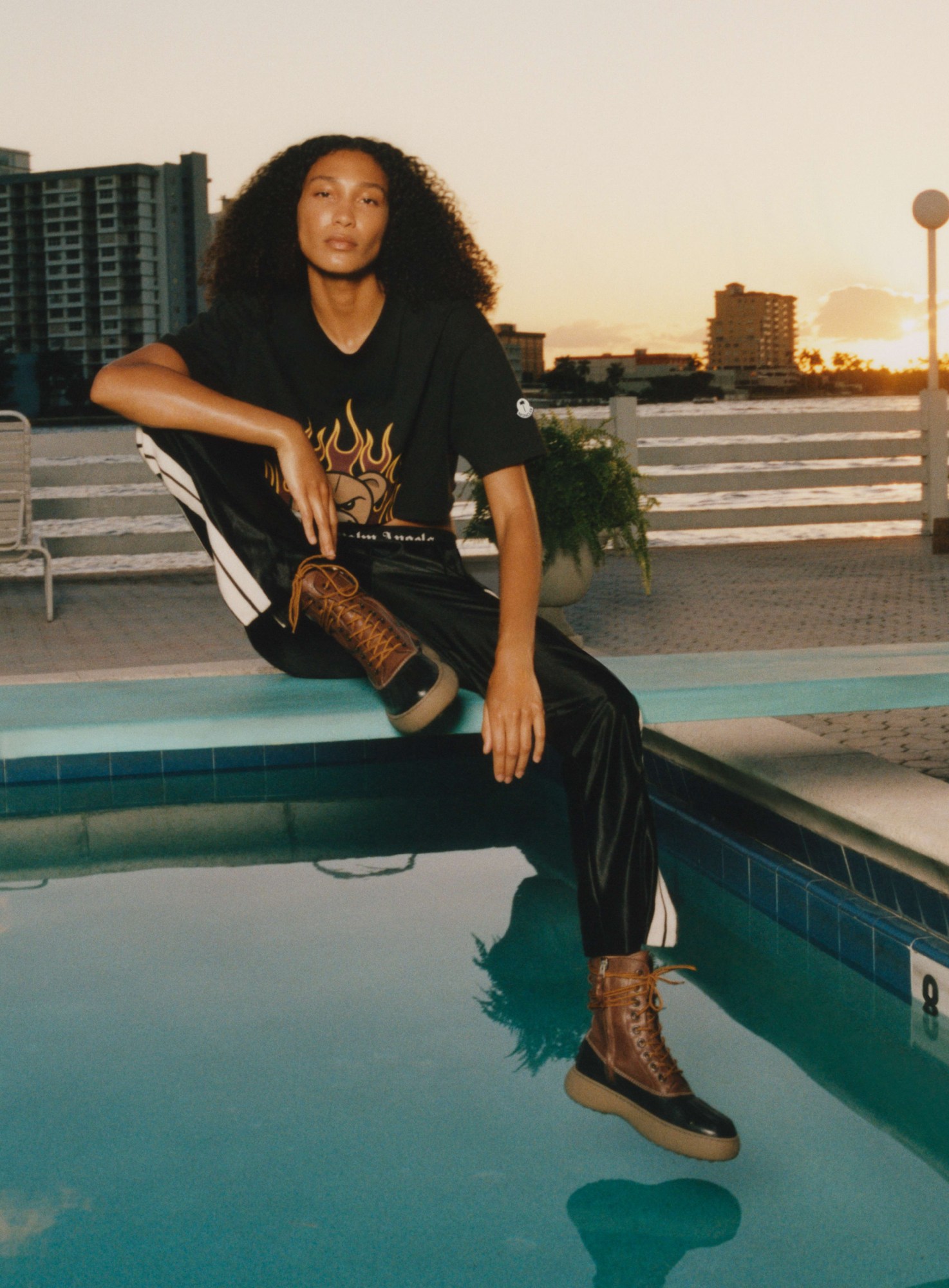 a model wearing tracksuit bottoms and leather boots sits on the side of a pool at sunset