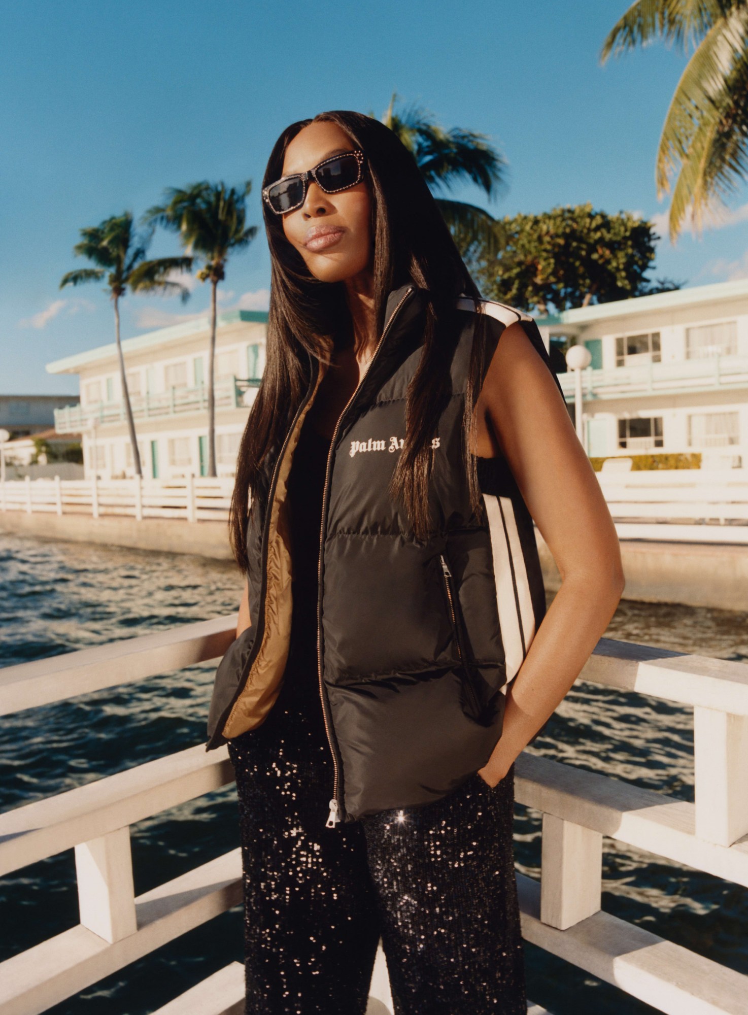 naomi campbell wearing a black puffer gilet with sequinned trousers, standing on a california boardwalk in the sun