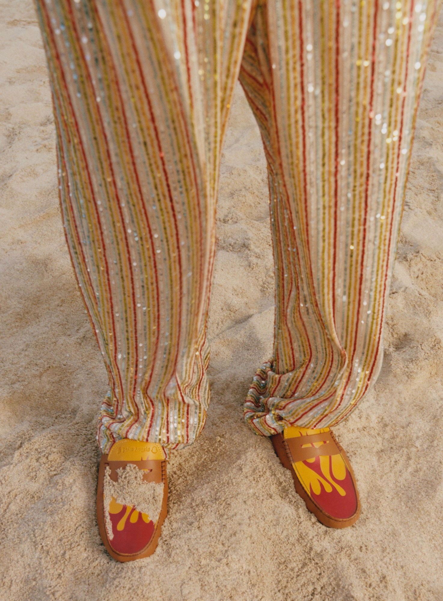 a close up of a models shiny beaded trousers and flame-painted deck shoes standing on a beach