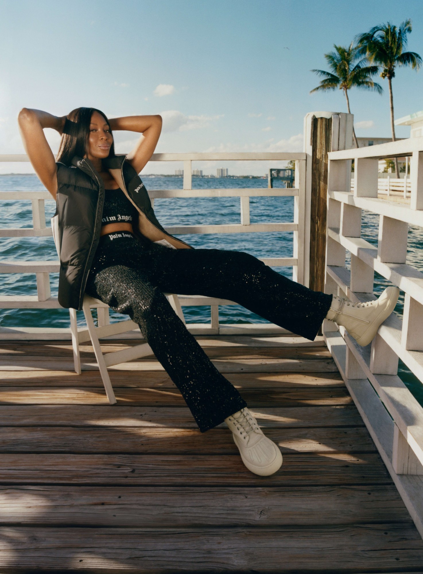 naomi cambell leans back in a chair on a sunny boardwalk, posing with her arms behind her head