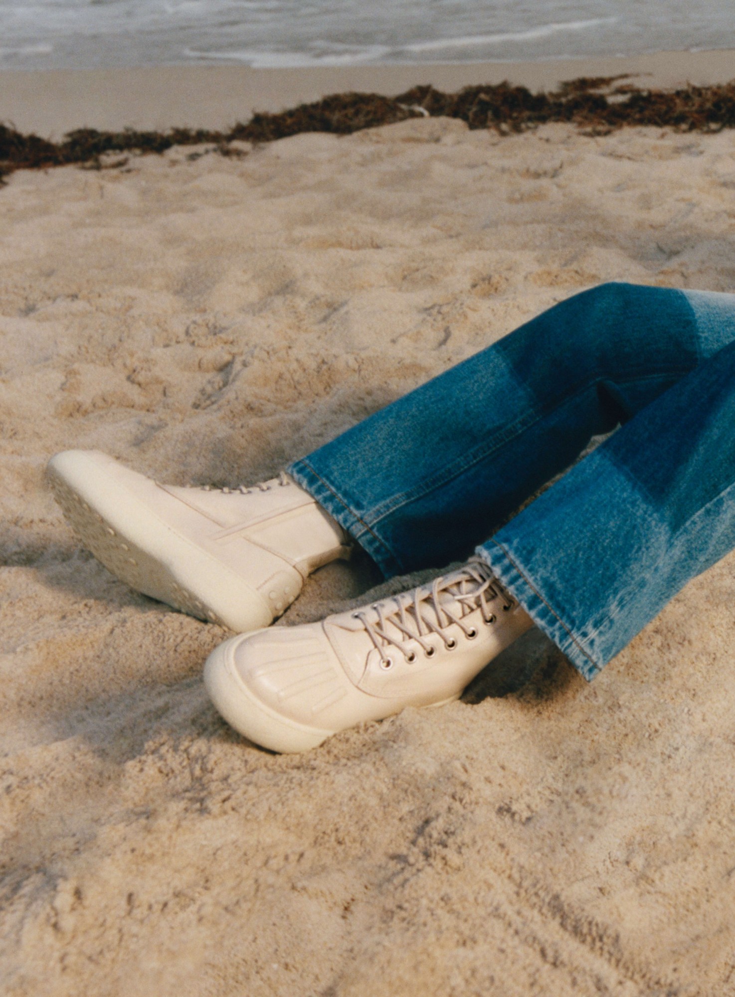 a close up of a model's white leather boots and jeans reclining on the beach