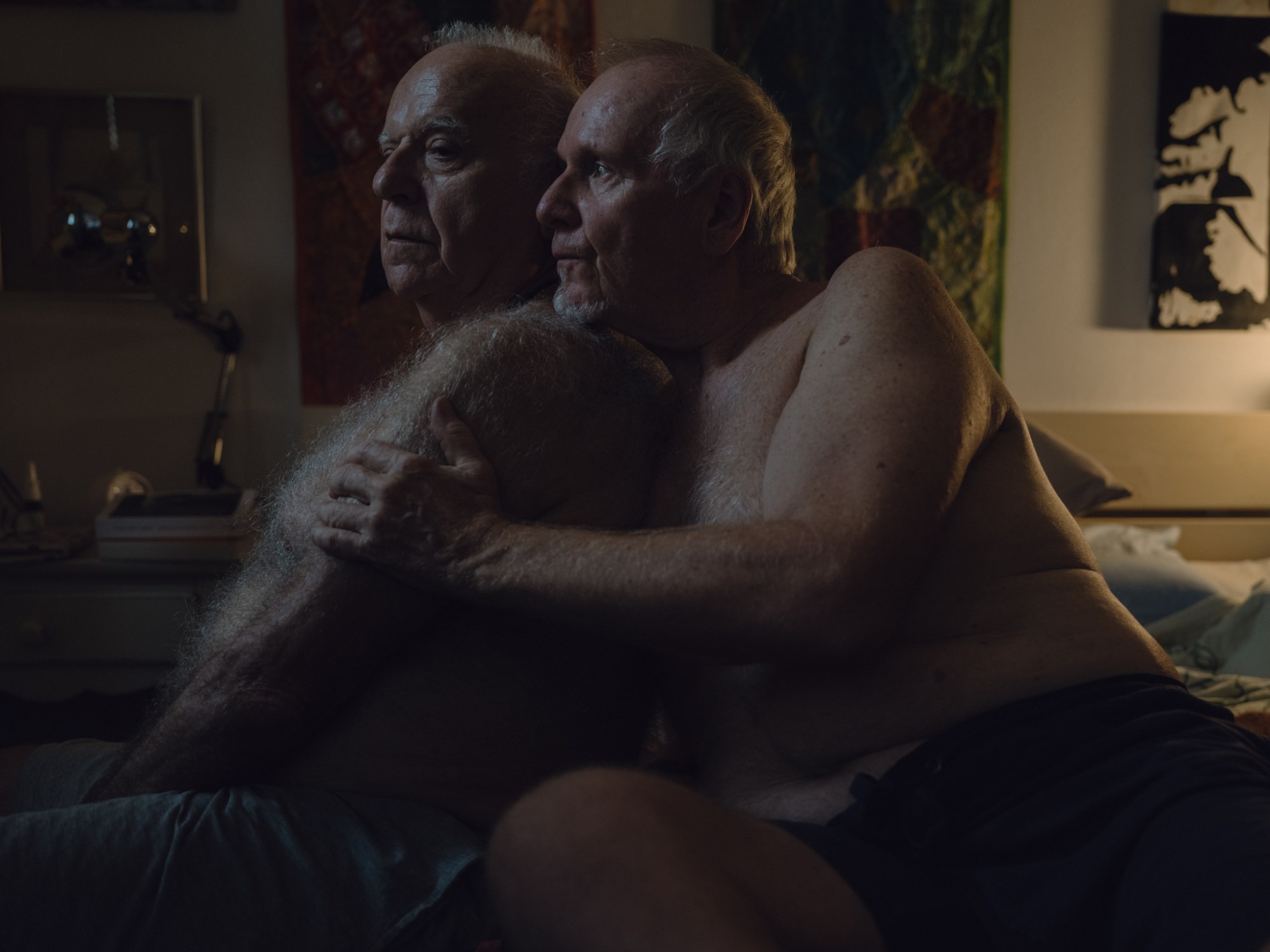 a topless older couple hold each other sitting on a bed, one rests his head on the other's shoulder