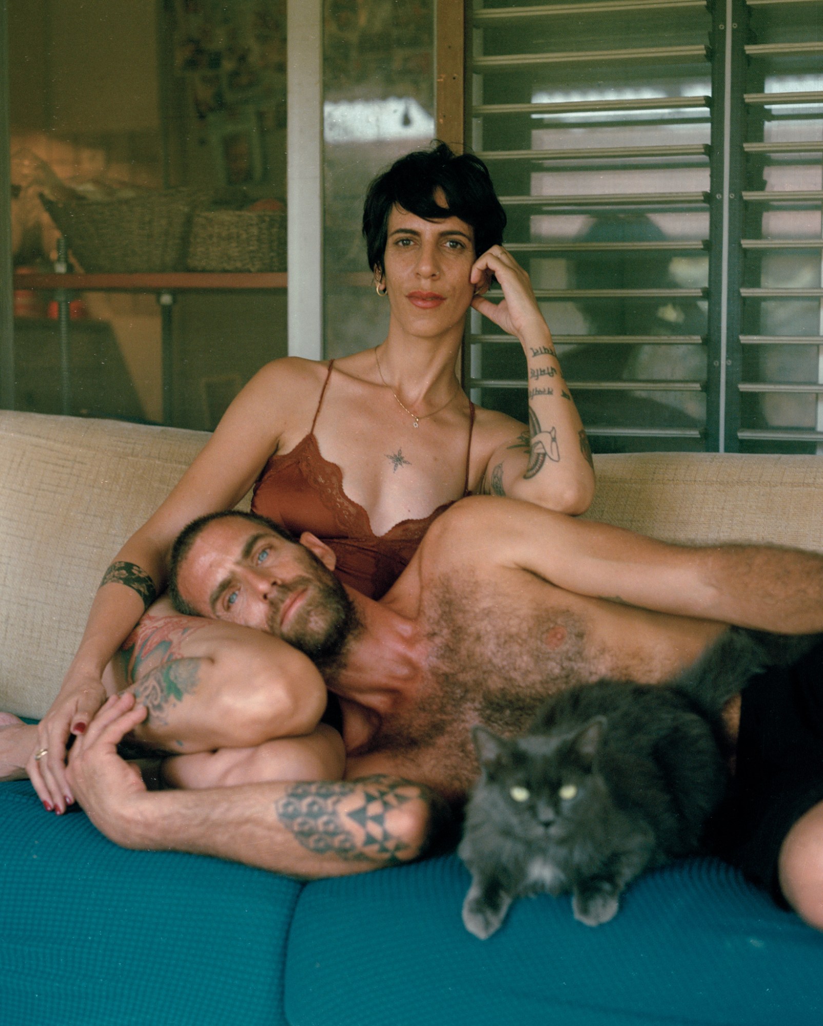 a couple sits on a sofa together (one with his head in the other's lap) with their cat