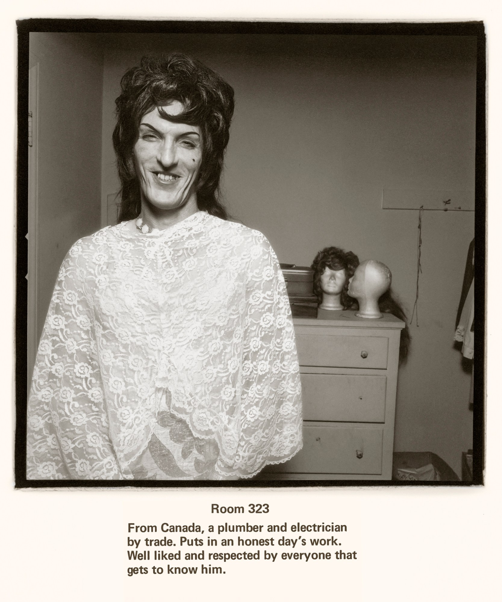 a person in a crocheted cape and dress looks into the camera smiling. they are wearing a wig . a dresser behind them has wig stands on it.