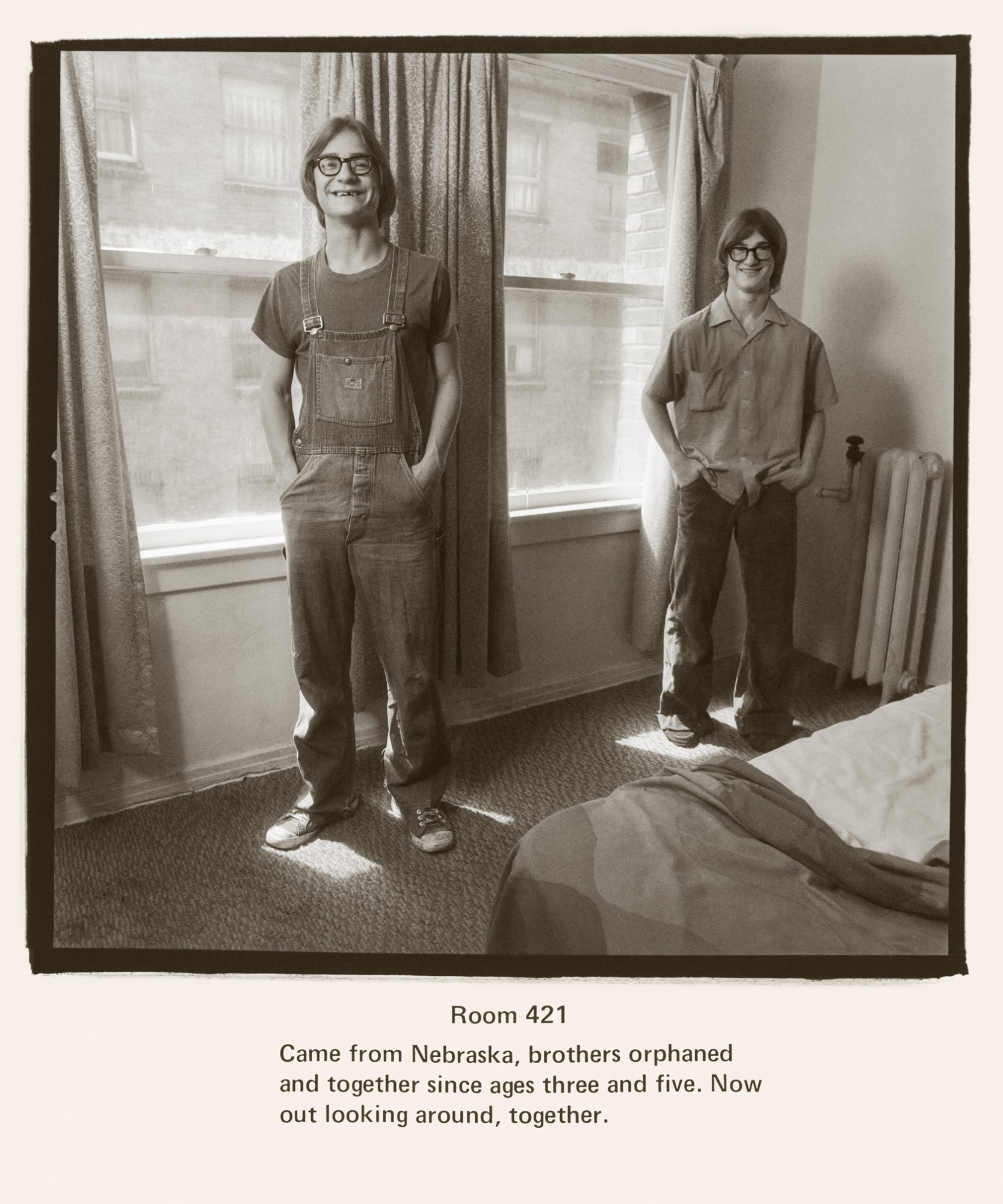 two brothers stand against two hotel room windows, one on the left wearing denim dungarees, one on the right in a camp collar shirt and slacks