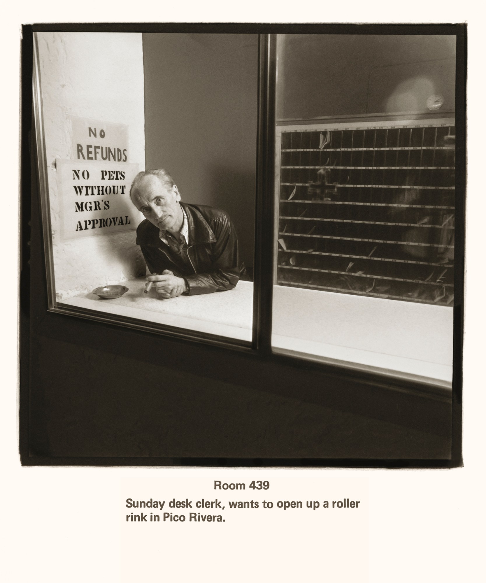 a man sits behind a hotel desk wearing a leather coat and shirt. he is balding and is smoking a cigarette. a sign behind him reads no refunds