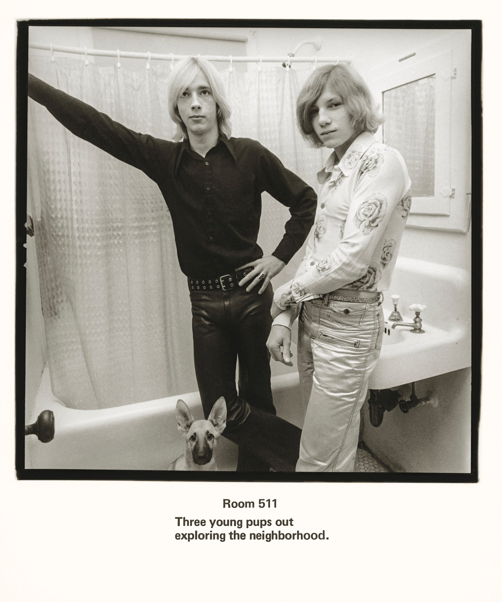 two young men pose in a pathroom. the one on the left is dressed in black with an open collar shirt and leather trousers. the one on the left is blonde wearing a floral shirt and jeans. a puppy dog is at their feet.