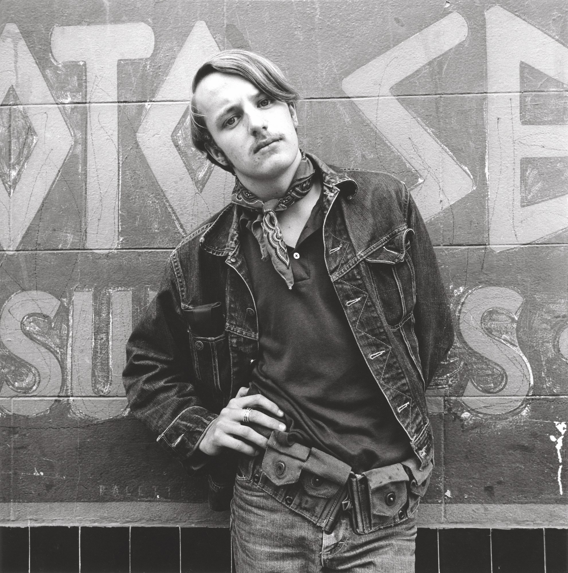 black-and-white image of a young man with a moustache, denim jacket and jeans with scarf around his neck