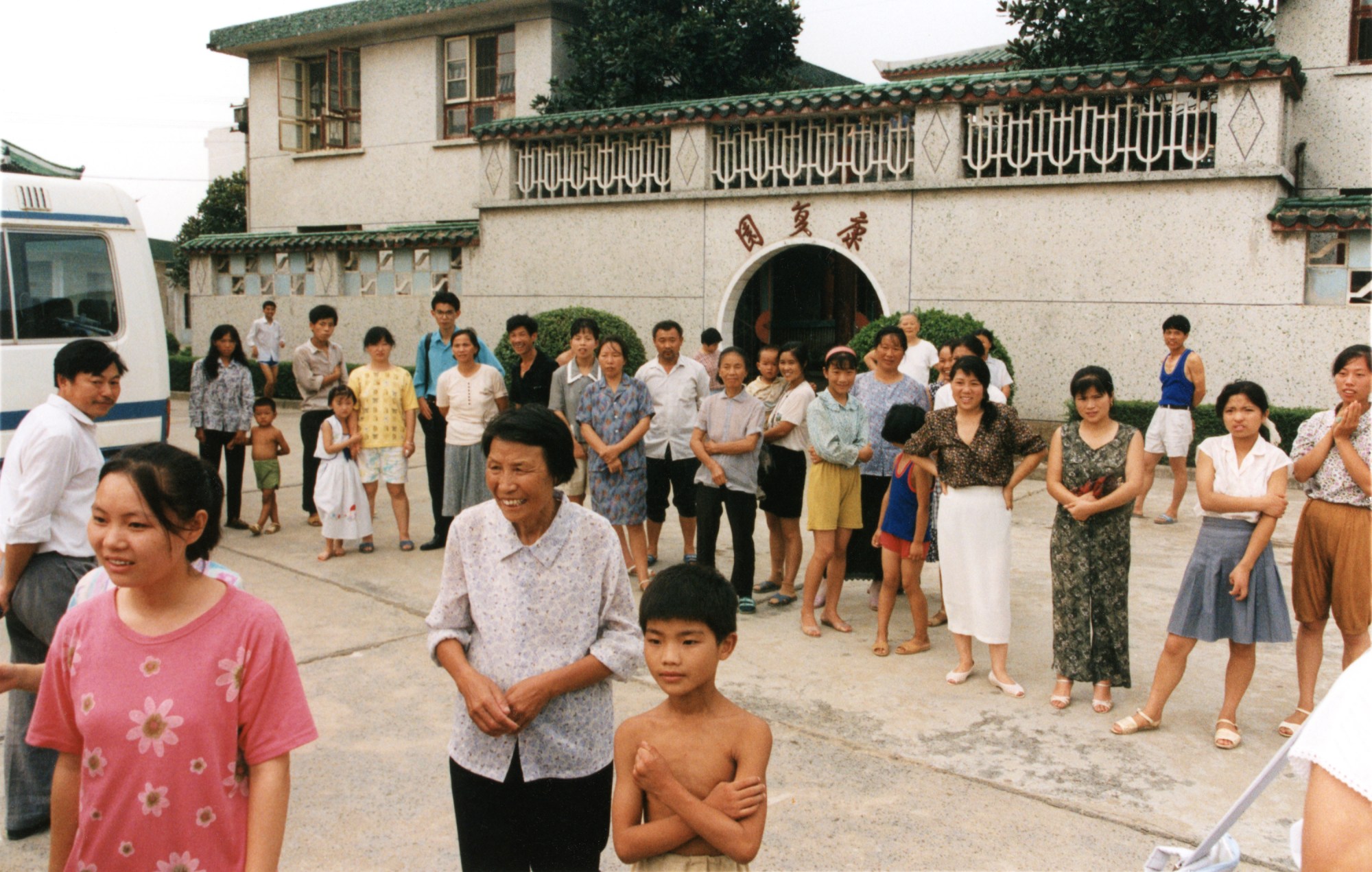 about 30 people of all ages stand around in front of a building, watching something. the photograph looks old