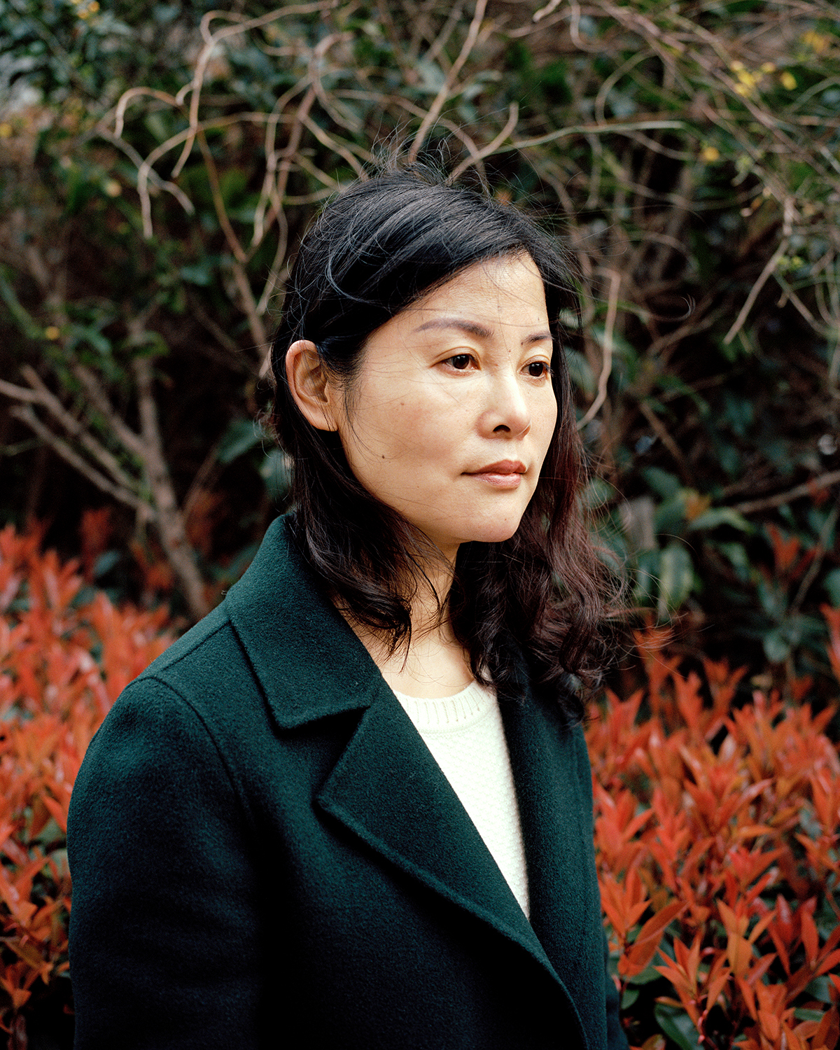 an attractive middle-aged chinese woman stands in front of a tree, looking off camera