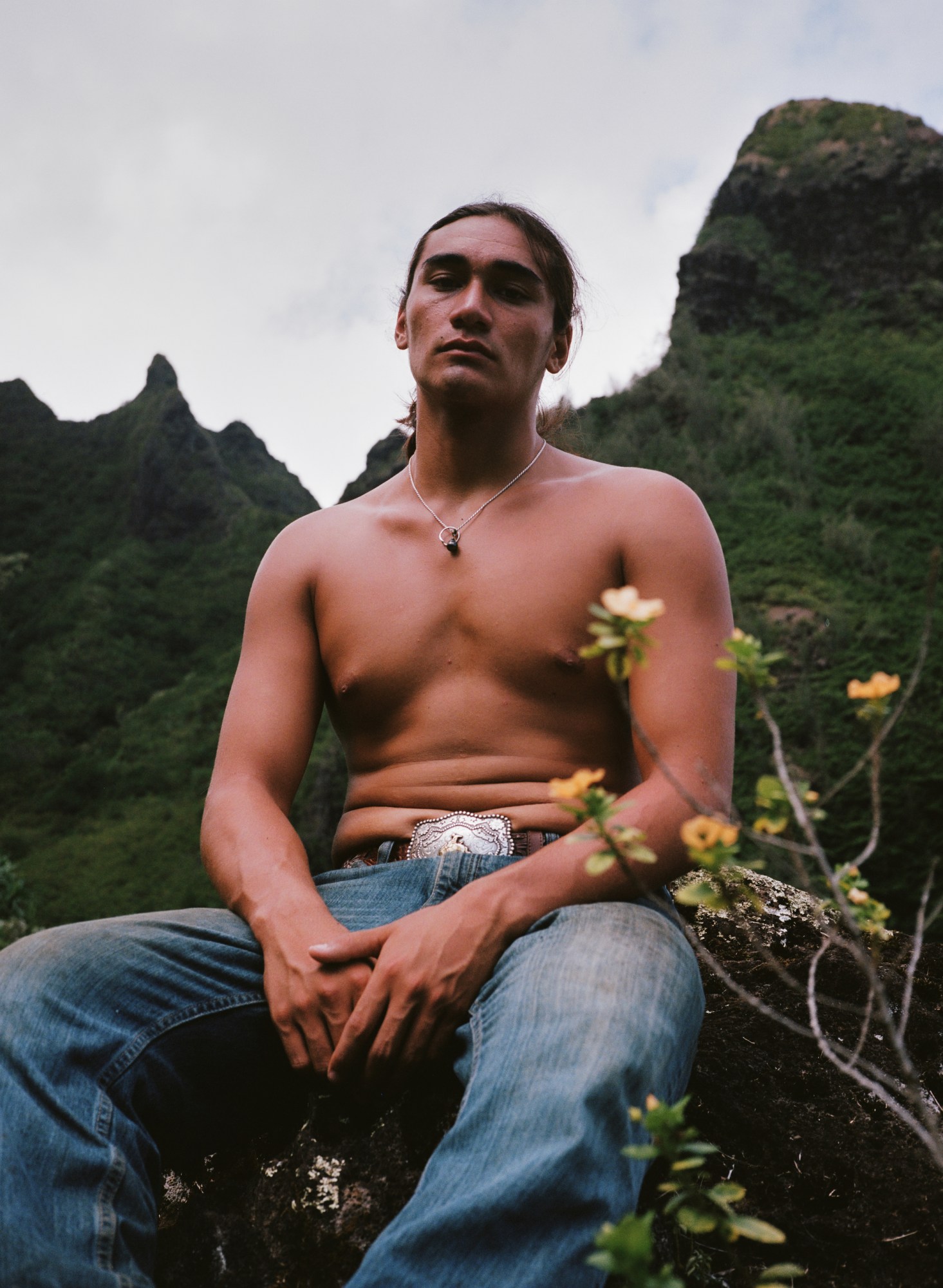 a man with his shirt off, jeans and a silver belt buckle pictured inthe grass