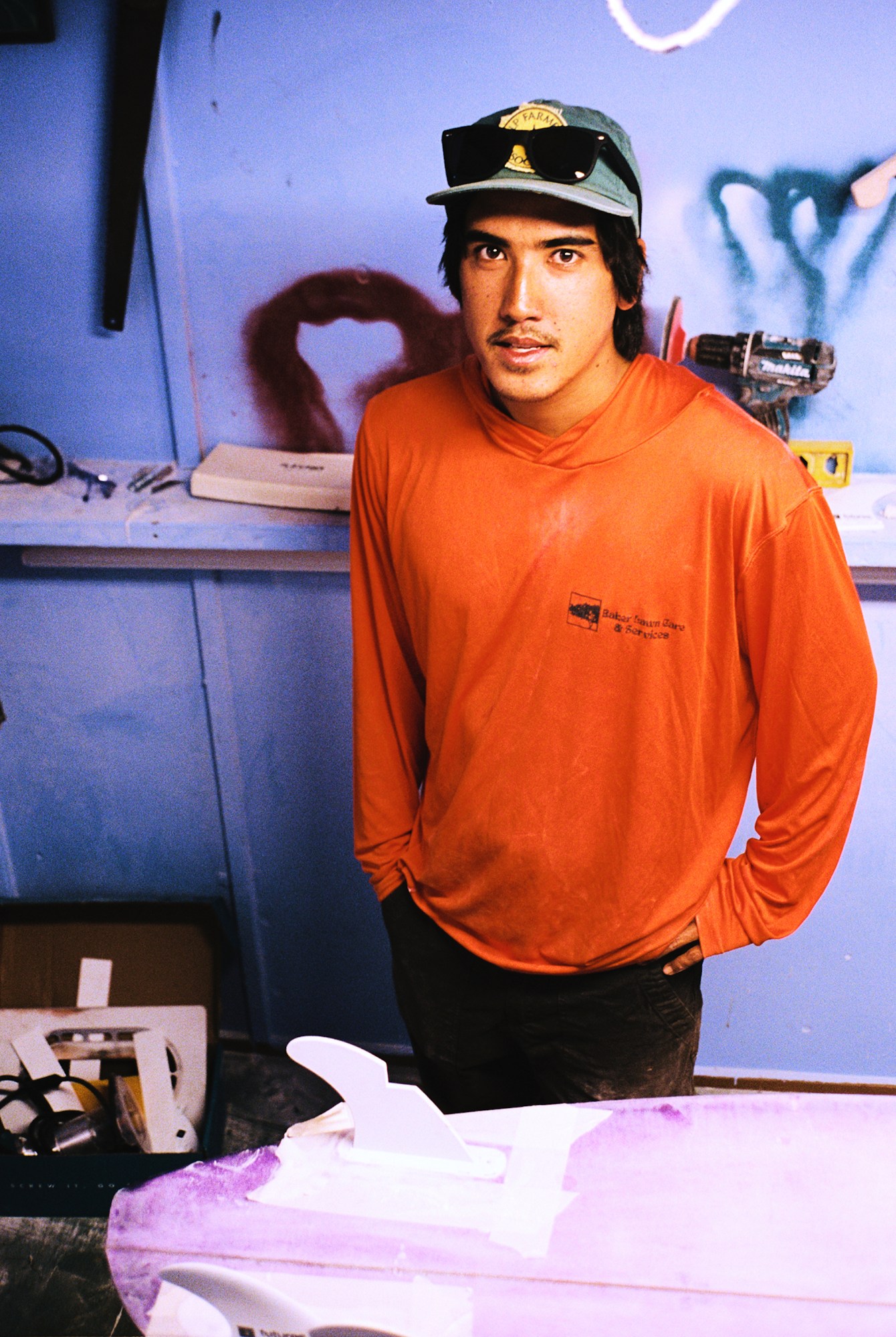 a guy in an orange shirt and hat standing in front of a surfboard