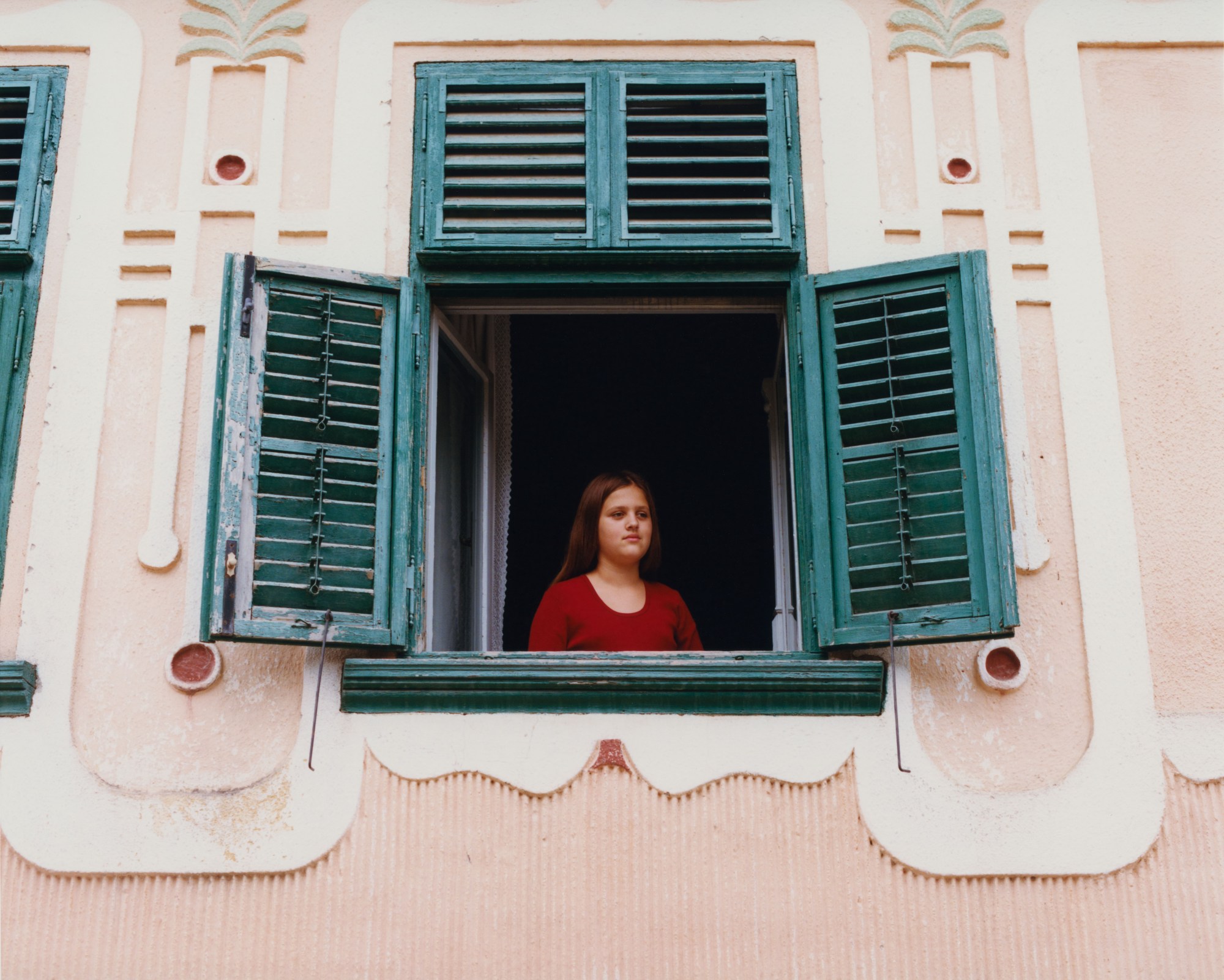 young girl looking out a green shuttered window framed by a pink wall