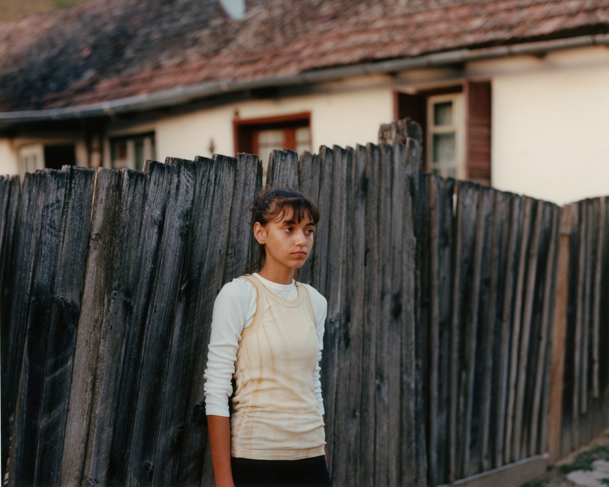 young girl in beige tank top and white long-sleeved top against a wooden fence