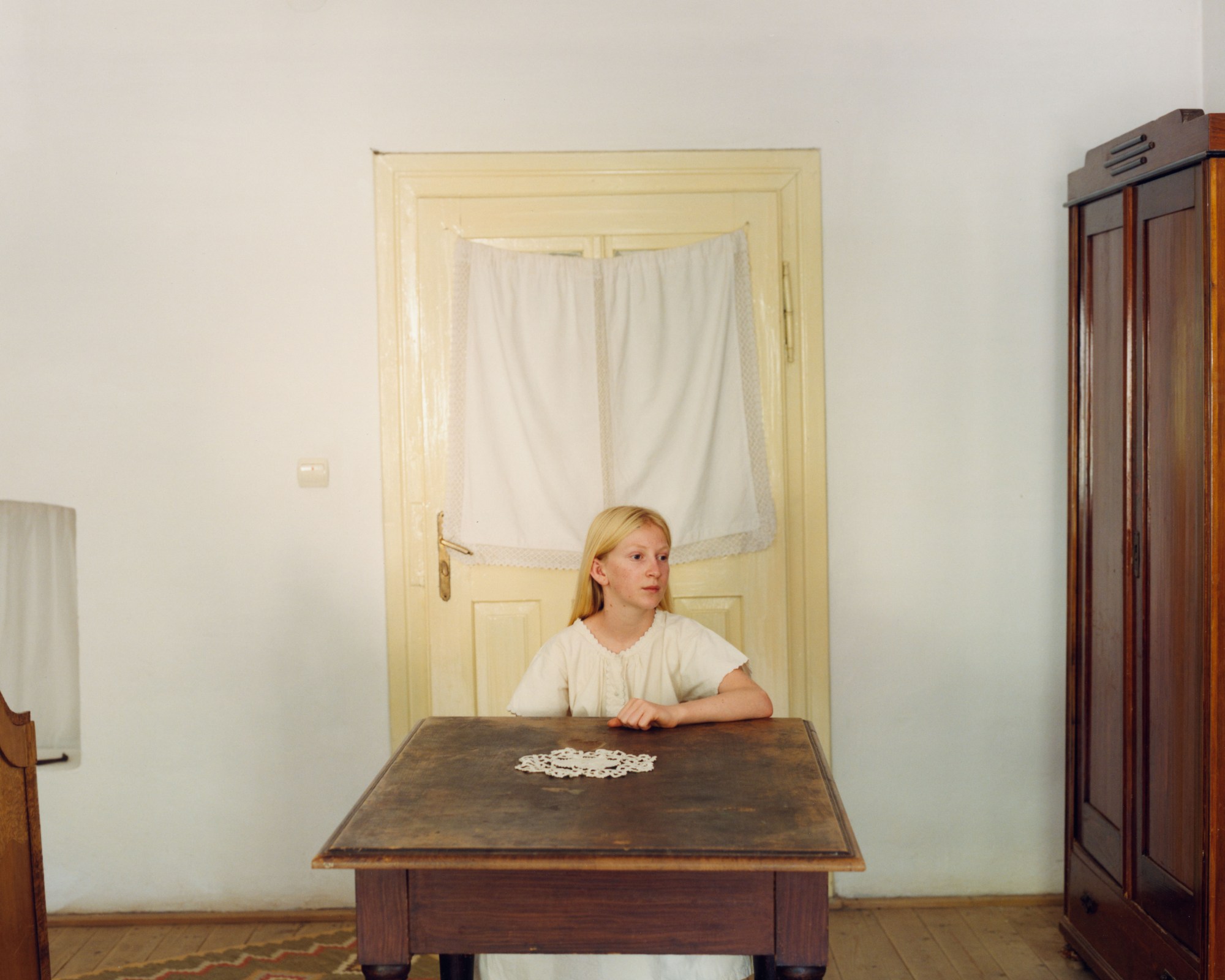 a young blonde girl sitting at a small wooden table in a white dress