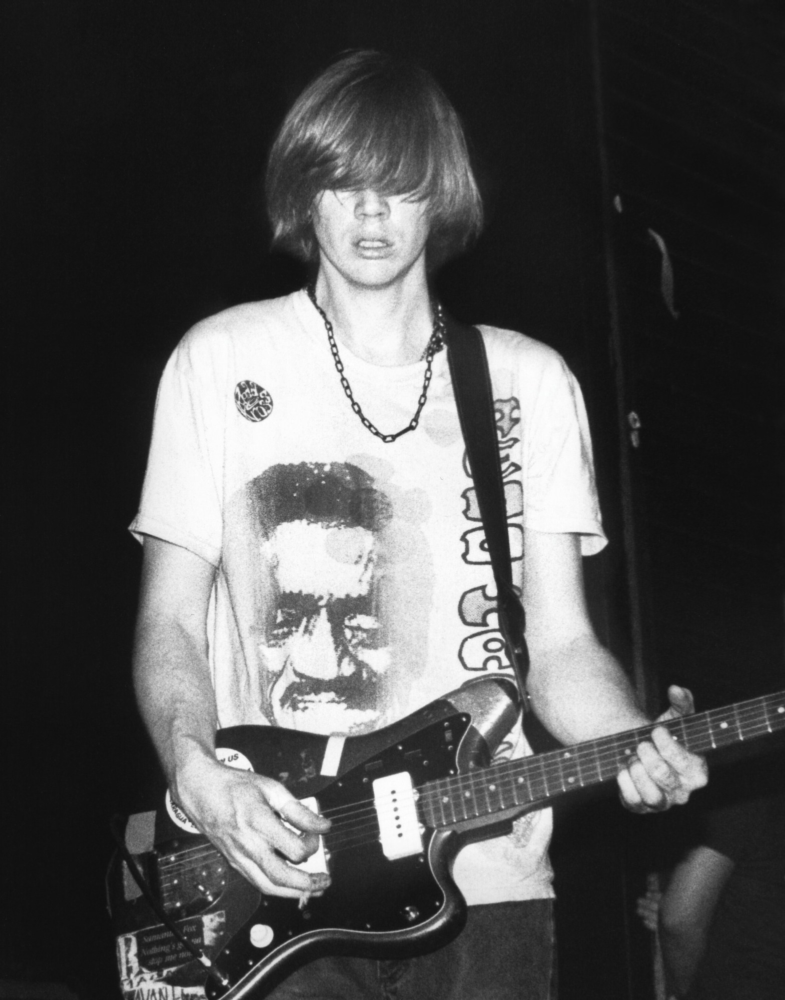 thurston moore of sonic youth, a young slim man with blonde hair that covers his eyes, plays guitar on stage. he wears a t-shirt with a man's face on it.