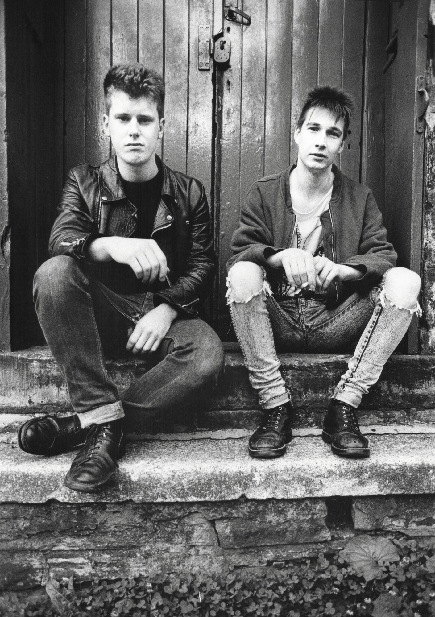 two men sit on the steps of a stone building, both wear jeans. one on the left is wearing a leather jacket, the one on the right wears a light bomber and a cut neck t-shirt underneath. they are white and have 80s hairstyles