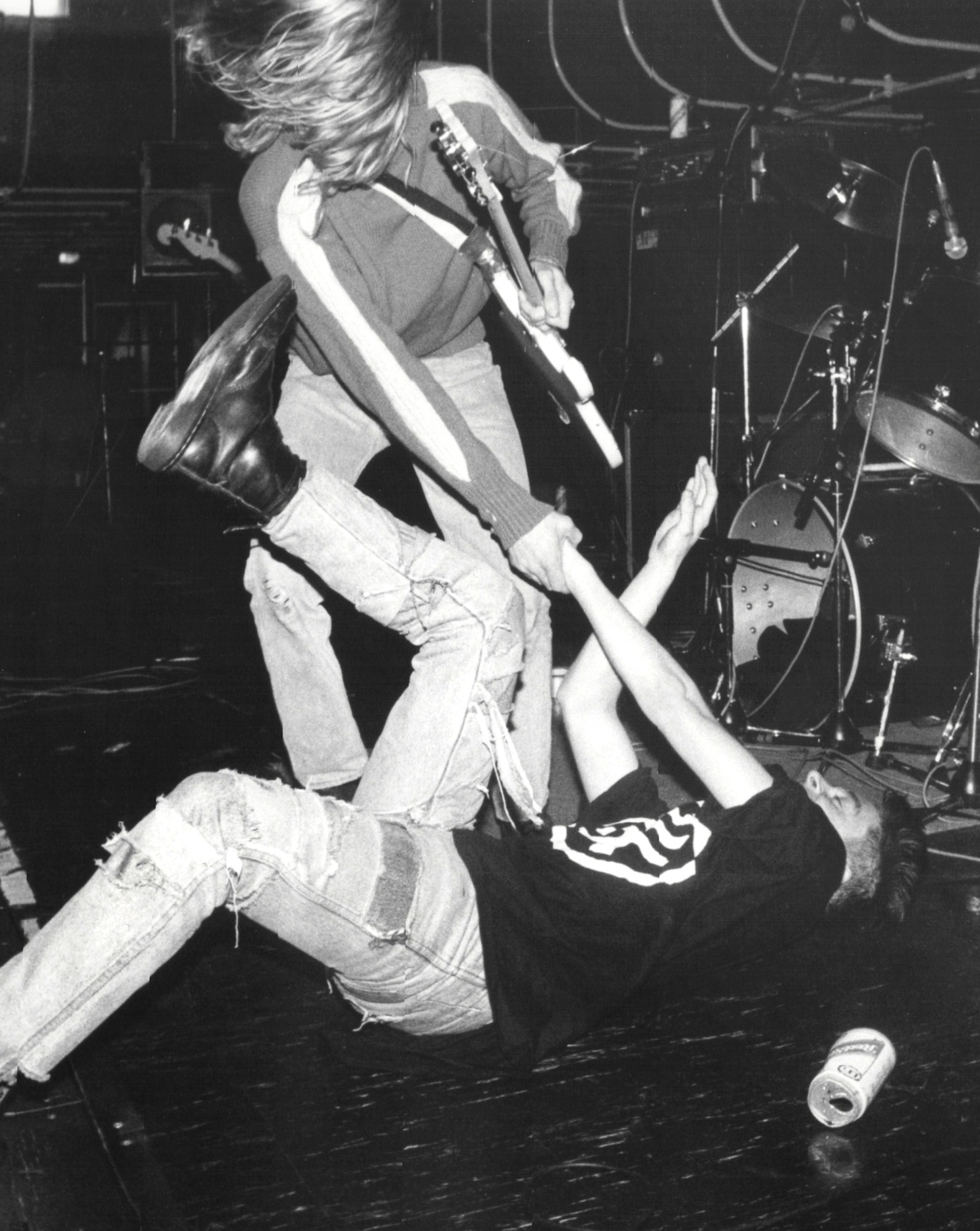 kurt cobain with long blonde hair wears a sweater with striped arms. he's holding his guitar in one hand and the hand of a fan who's fallen over on stage. the fan is wearing distressed denim jeans and a black logo t shirt.