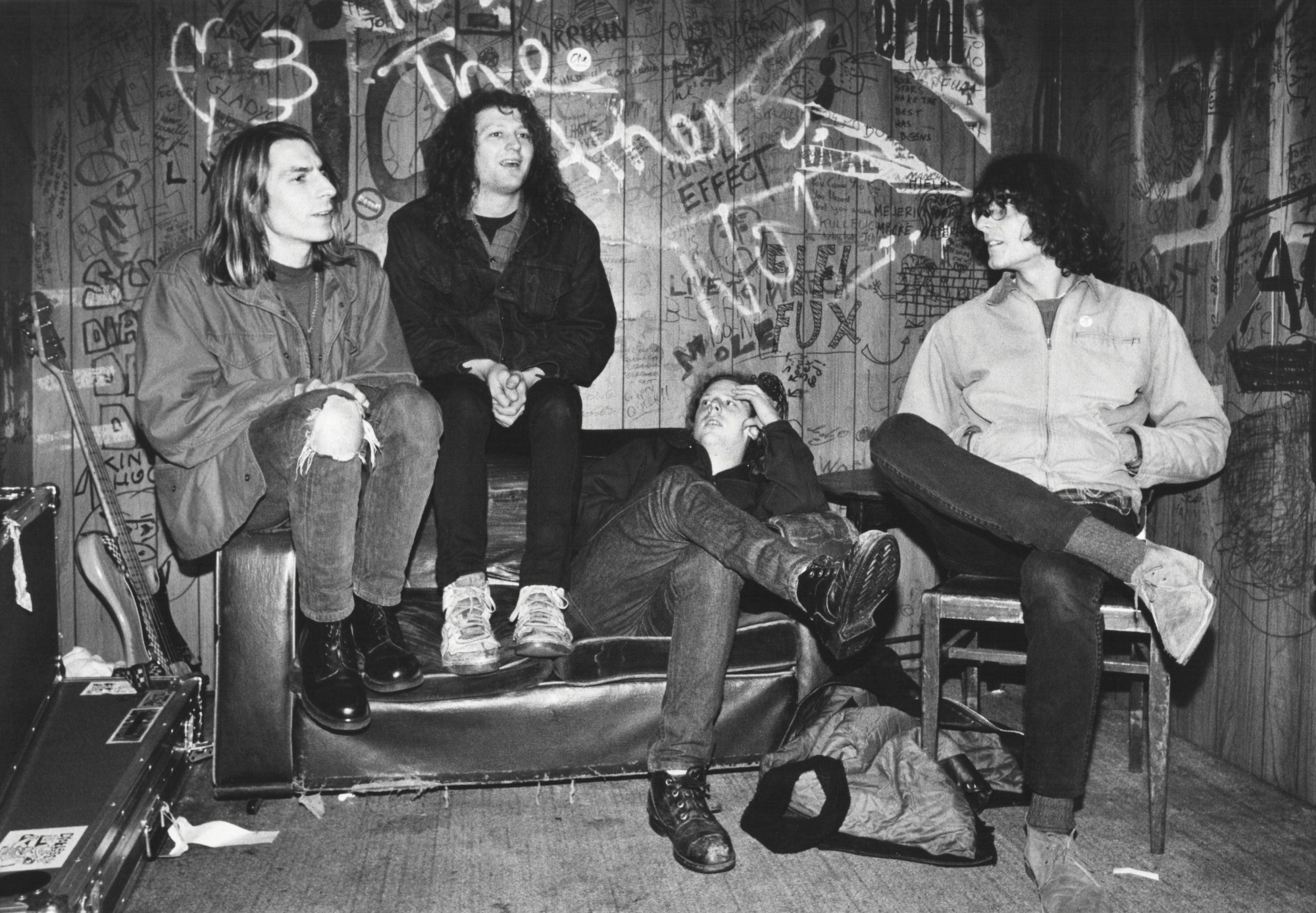 a band sits on a frayed leather couch against a wooden wall spraypainted with grafitti. they wear skinny denim jeans and have long shoulder length hair. they look like they're in conversation with each other.